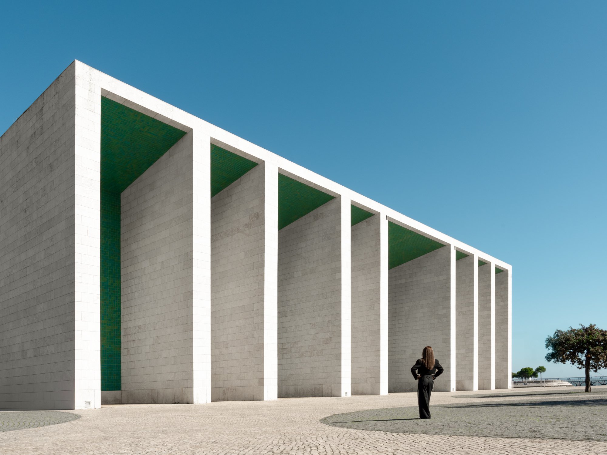 A woman in black standing with her hands on her hips, looking up at a modern white building with tall vertical columns and green interior walls against a clear blue sky with a few trees nearby.