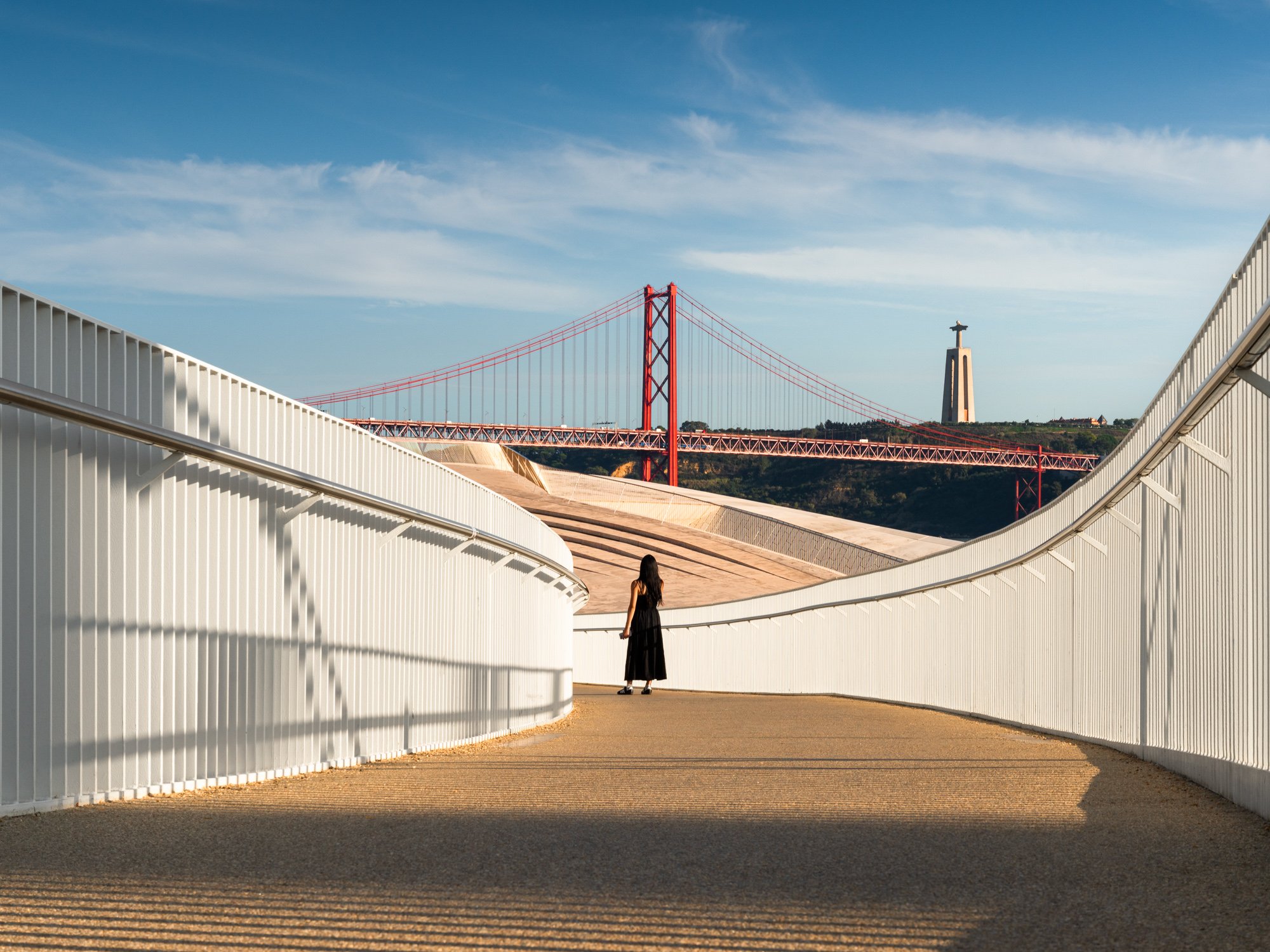 Woman walking on a modern pathway with white railings, featuring the Golden Gate Bridge and Christ the Redeemer statue in the background under a blue sky.