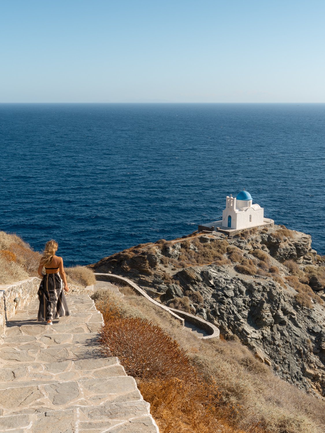 A woman with long blonde hair walking down a stone pathway on a rocky hillside towards a white building with a blue dome near the sea.