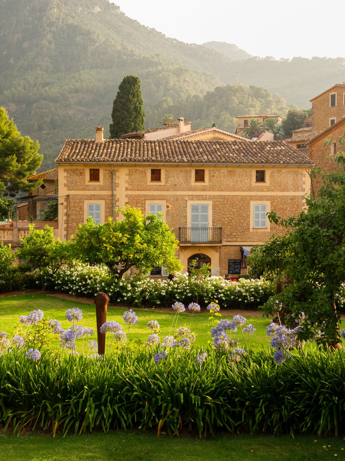 A stone building with a tiled roof and white shutters, surrounded by greenery and flowering bushes, with a mountain in the background.