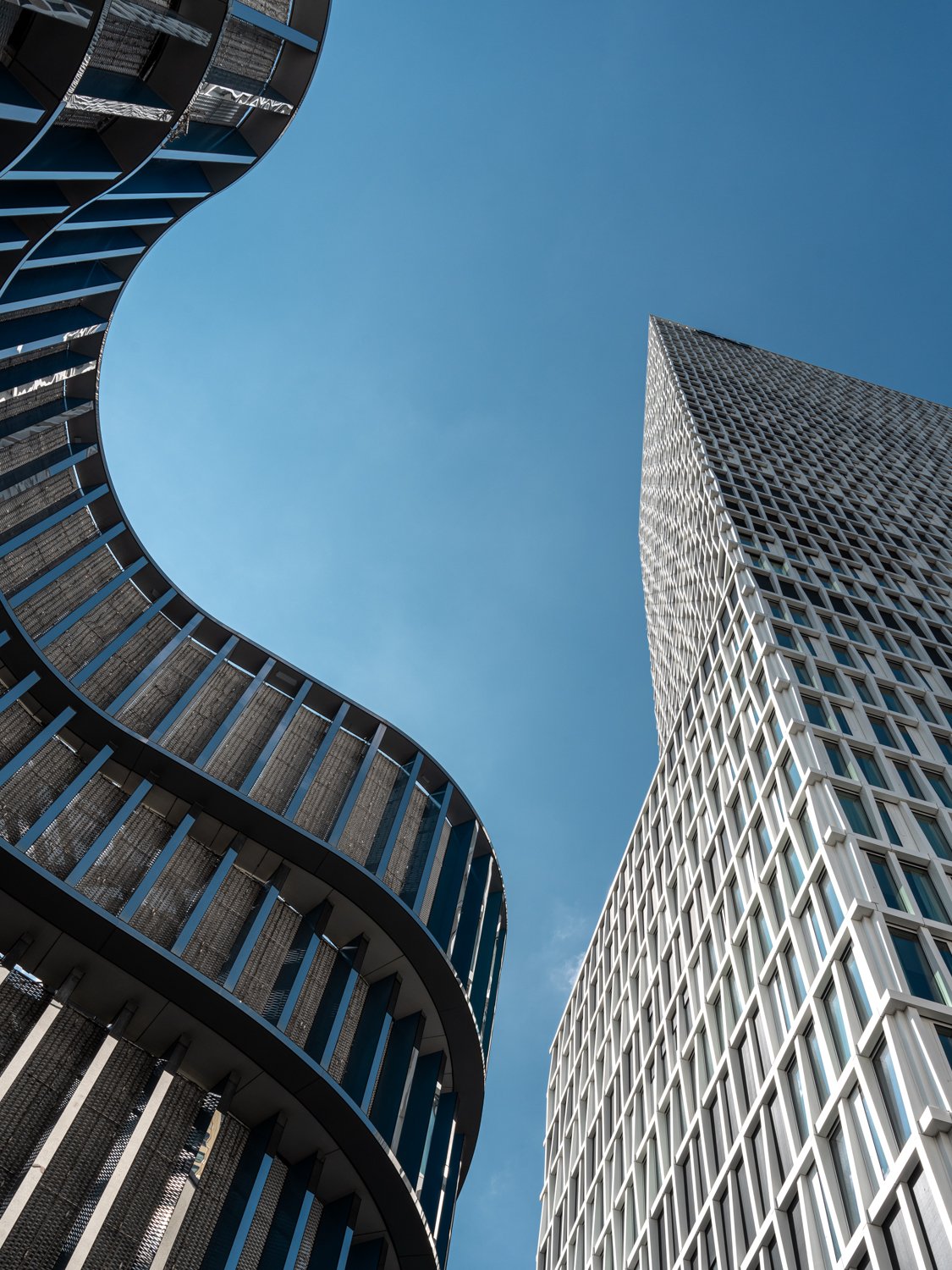 Looking up at modern skyscrapers with glass windows and unique architectural designs against a blue sky.