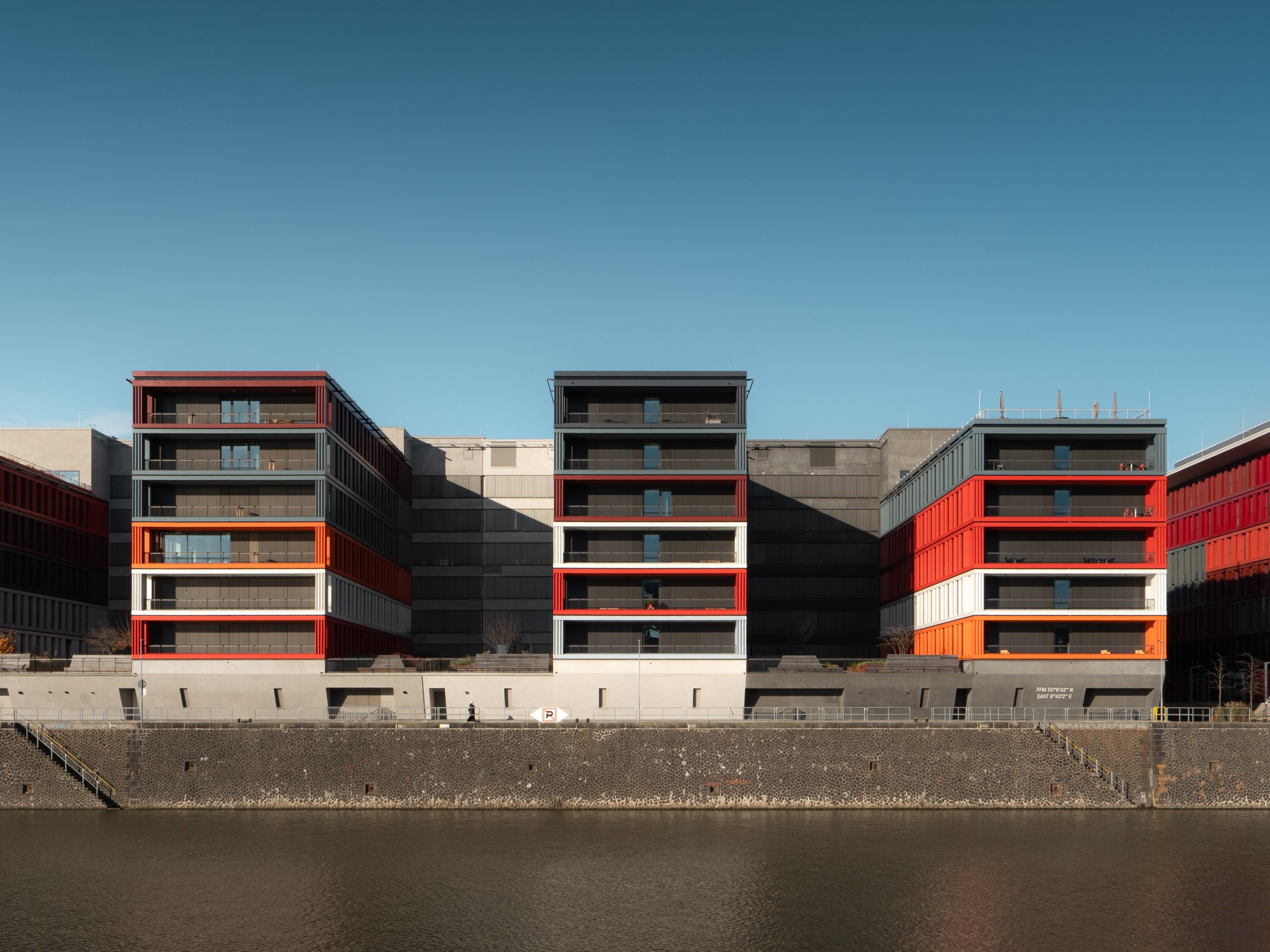 Modern multi-story apartment buildings with red, black, gray, and white exterior accents, situated along a waterway and under a clear blue sky.