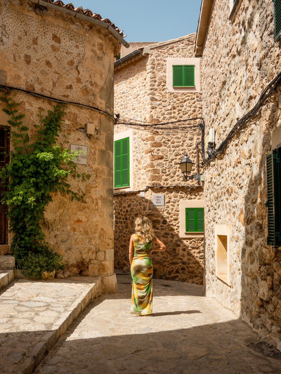 A woman in a colorful, flowing dress walking through a narrow, stone-paved alley with old stone buildings and green window shutters on a sunny day.