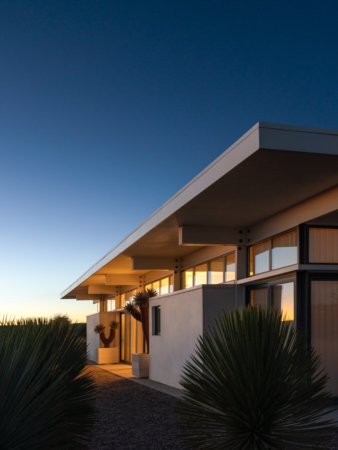 Modern house with large glass windows and white walls, surrounded by desert plants, during sunset with a clear sky.