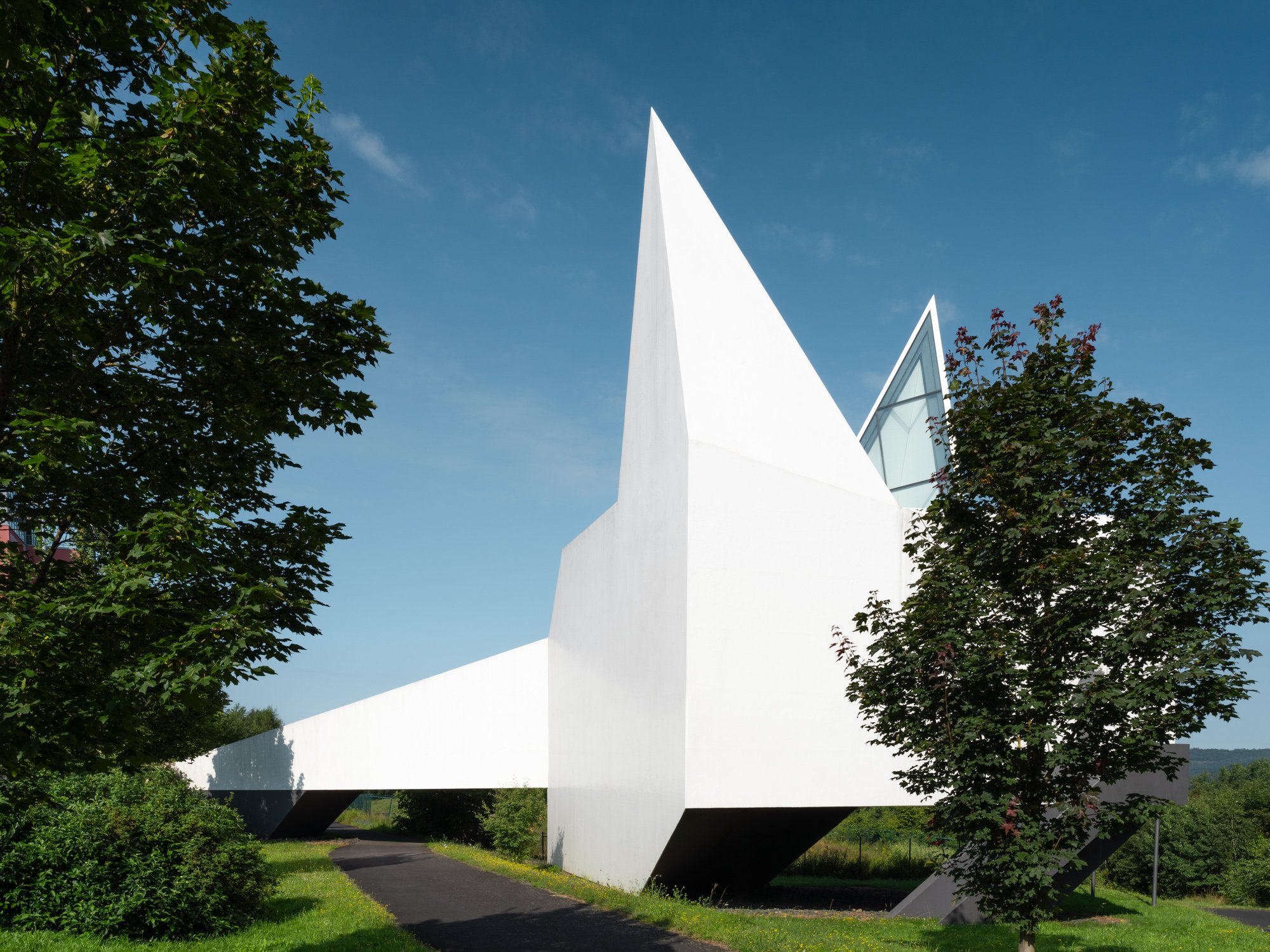 Modern white building with sharp angular design, surrounded by green trees and under a clear blue sky.