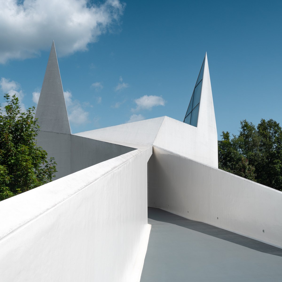 Modern white architectural structure with angular design and sharp pointed spires, set against a blue sky with scattered clouds.
