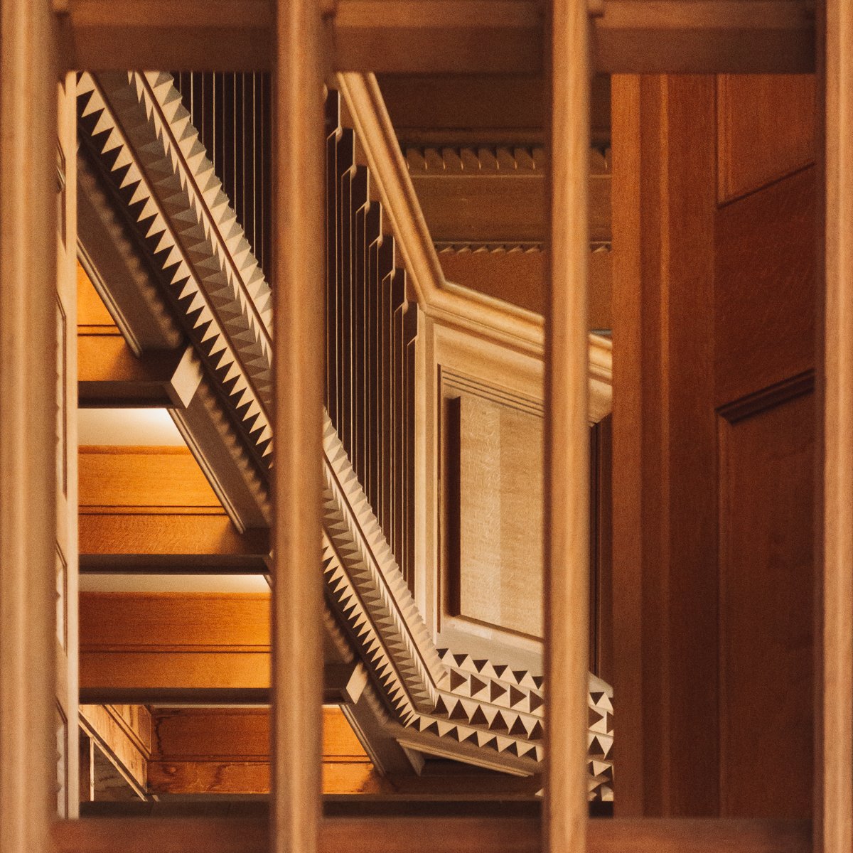 Interior view of a wooden staircase seen through a banister with vertical bars, looking upwards from the bottom of the staircase.