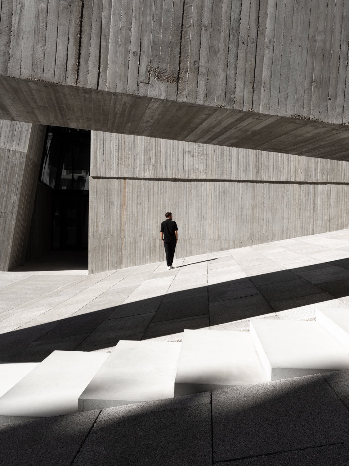 A person walking on a modern, minimalist concrete building exterior with geometric architectural design, creating shadow patterns.