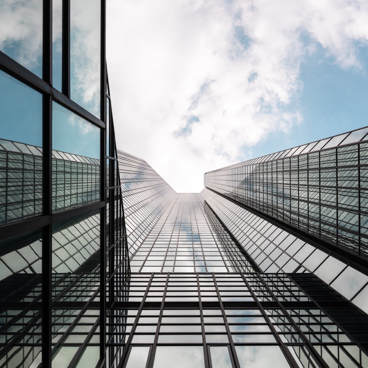 Looking up at a tall modern glass skyscraper with reflections of the sky and clouds, from street level.