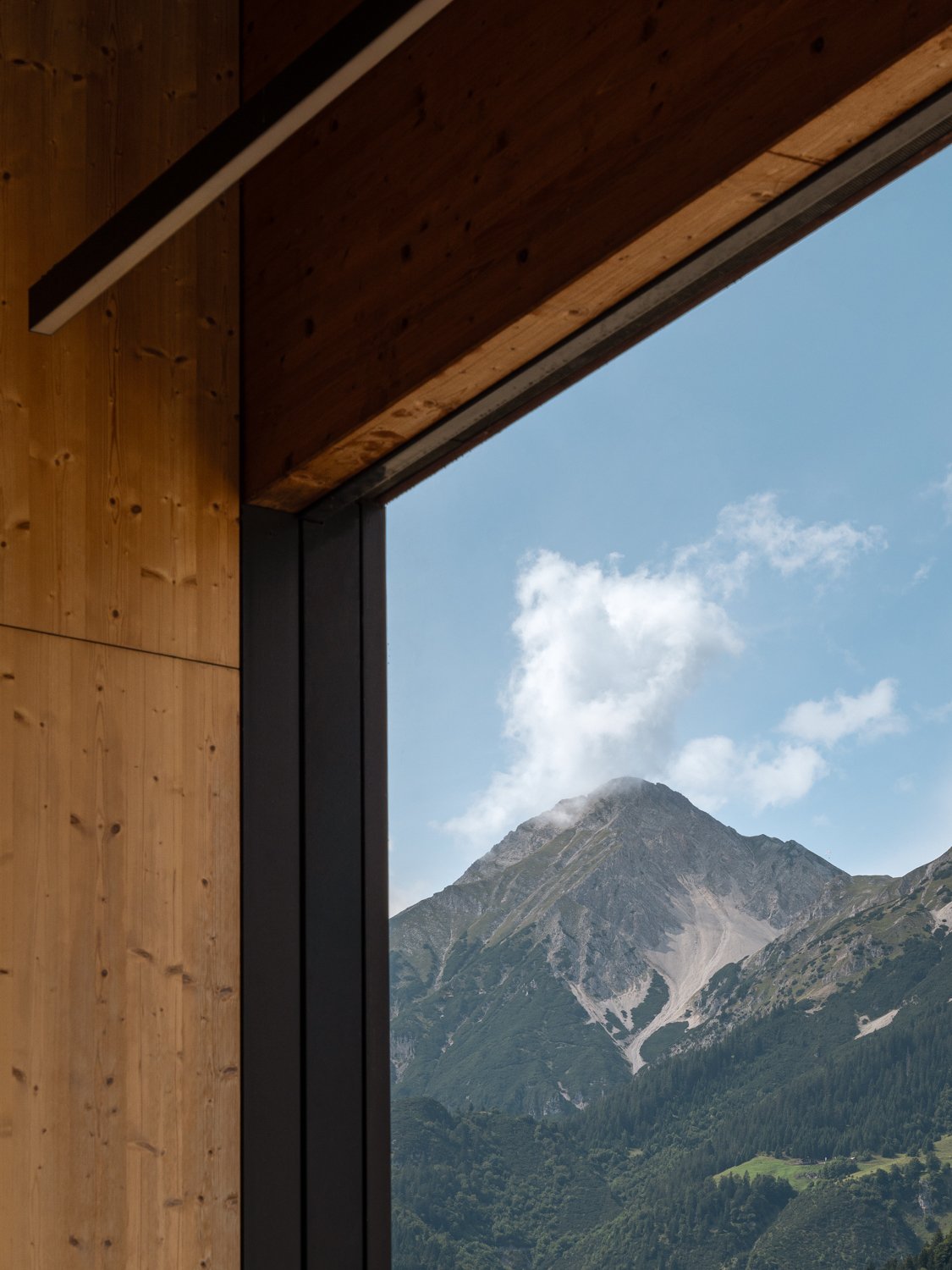 View of a mountain with a partly cloudy sky seen through a large window in a wooden room.