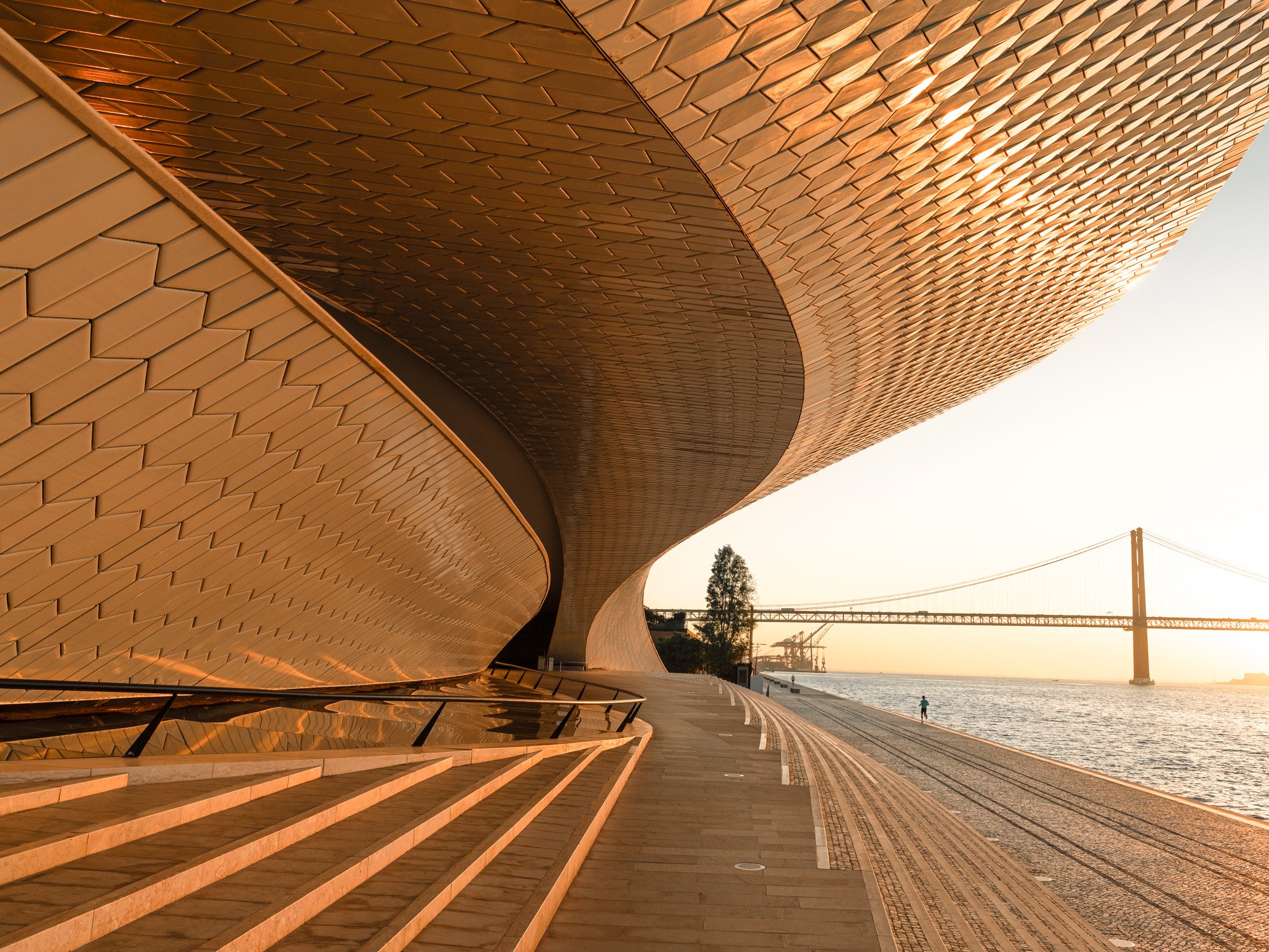 Modern architectural building with a curved design and textured copper-colored exterior, situated along a waterfront with train tracks, a bridge, and a person walking near the water at sunset.