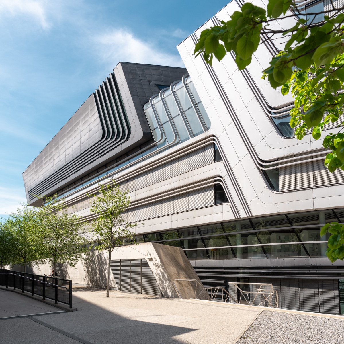 Modern multi-story building with curved architectural design, glass windows, and black accents, surrounded by trees and a sidewalk under a blue sky.
