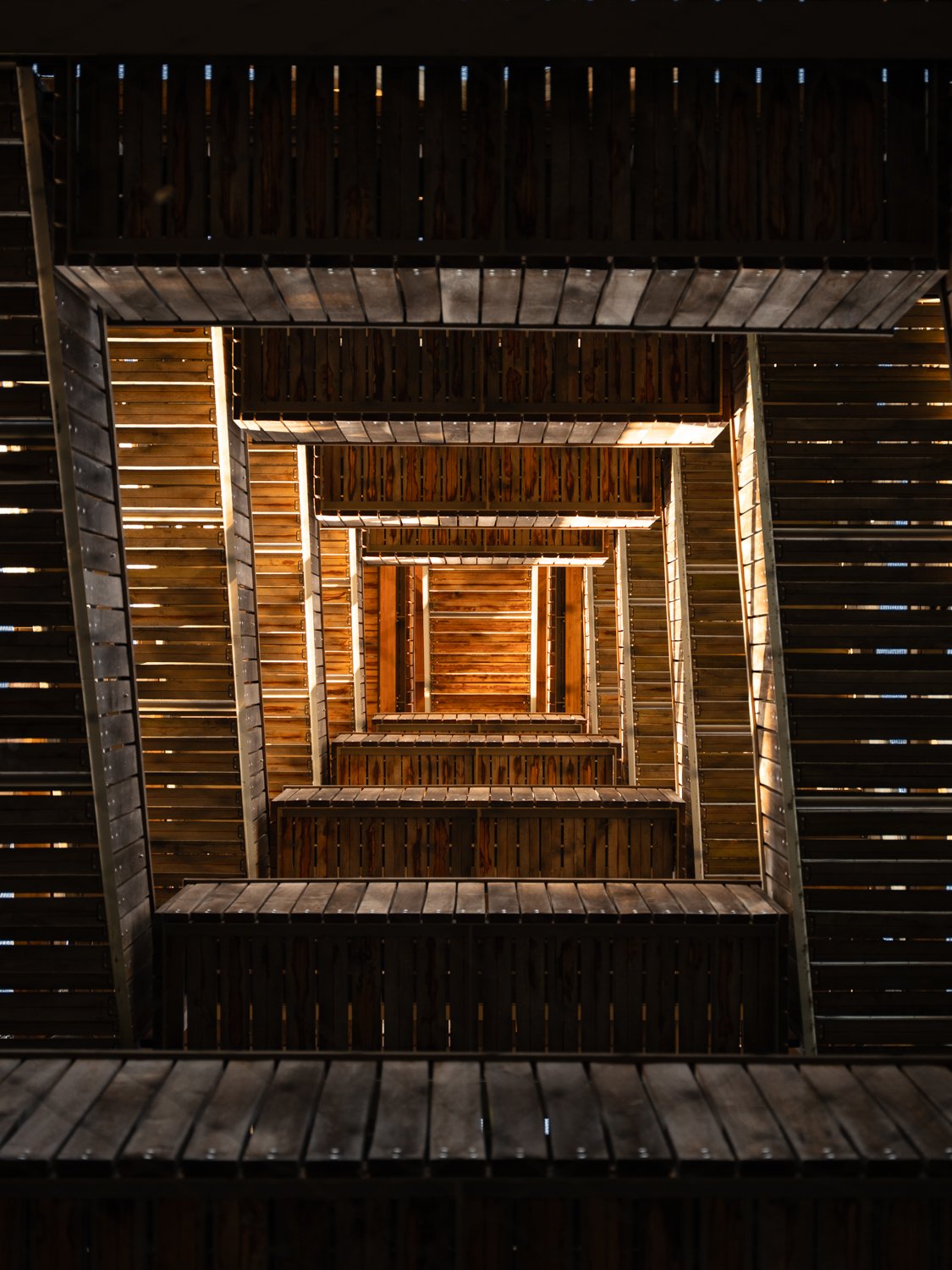 Looking up at a wooden spiral staircase with wooden railings and steps, illuminated by natural light coming through gaps in the wood.
