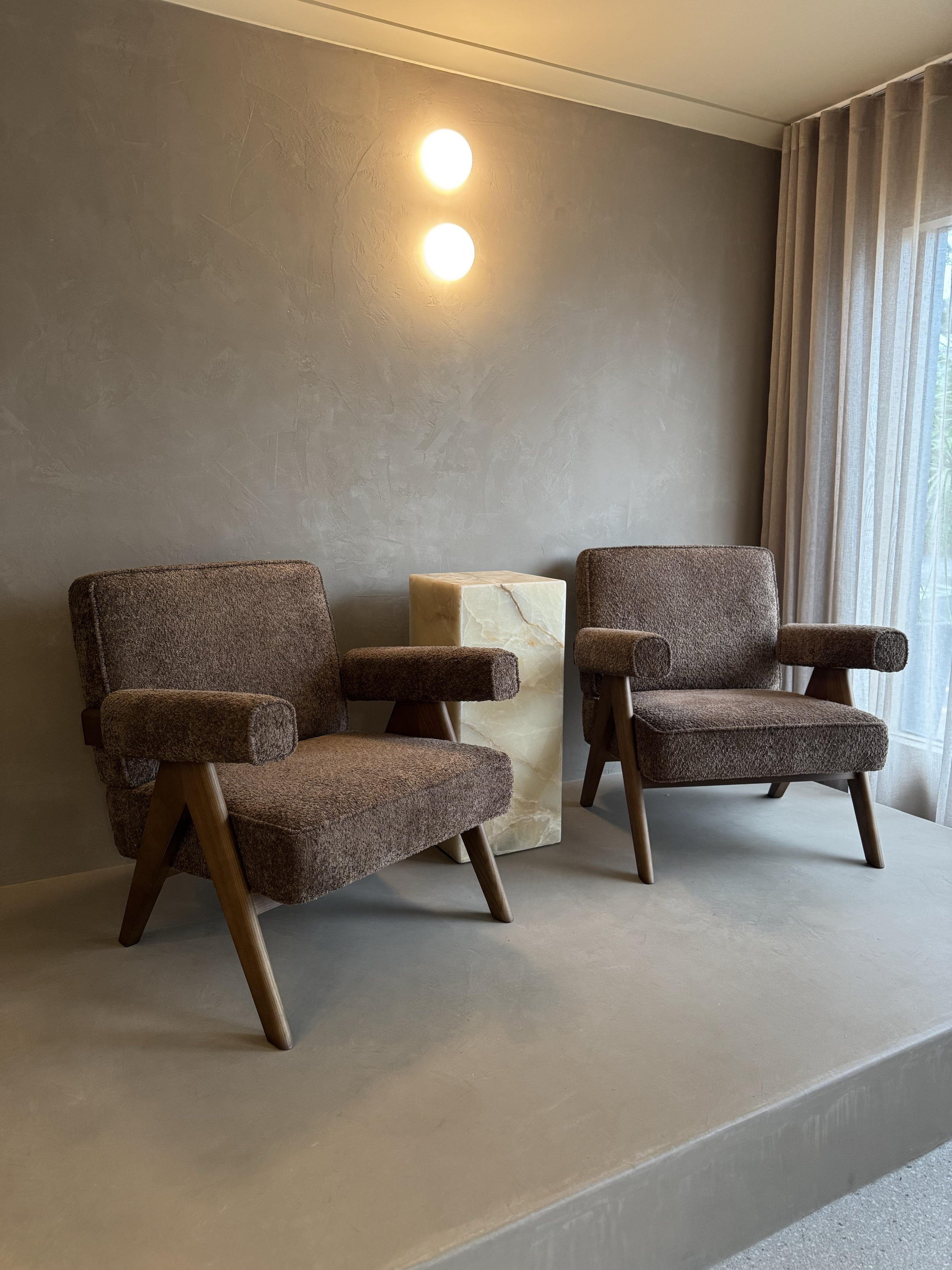 Two vintage-style brown upholstered armchairs with wooden legs, a marble side table between them, a beige textured wall with two round wall lights, and a window with sheer curtains on the right.