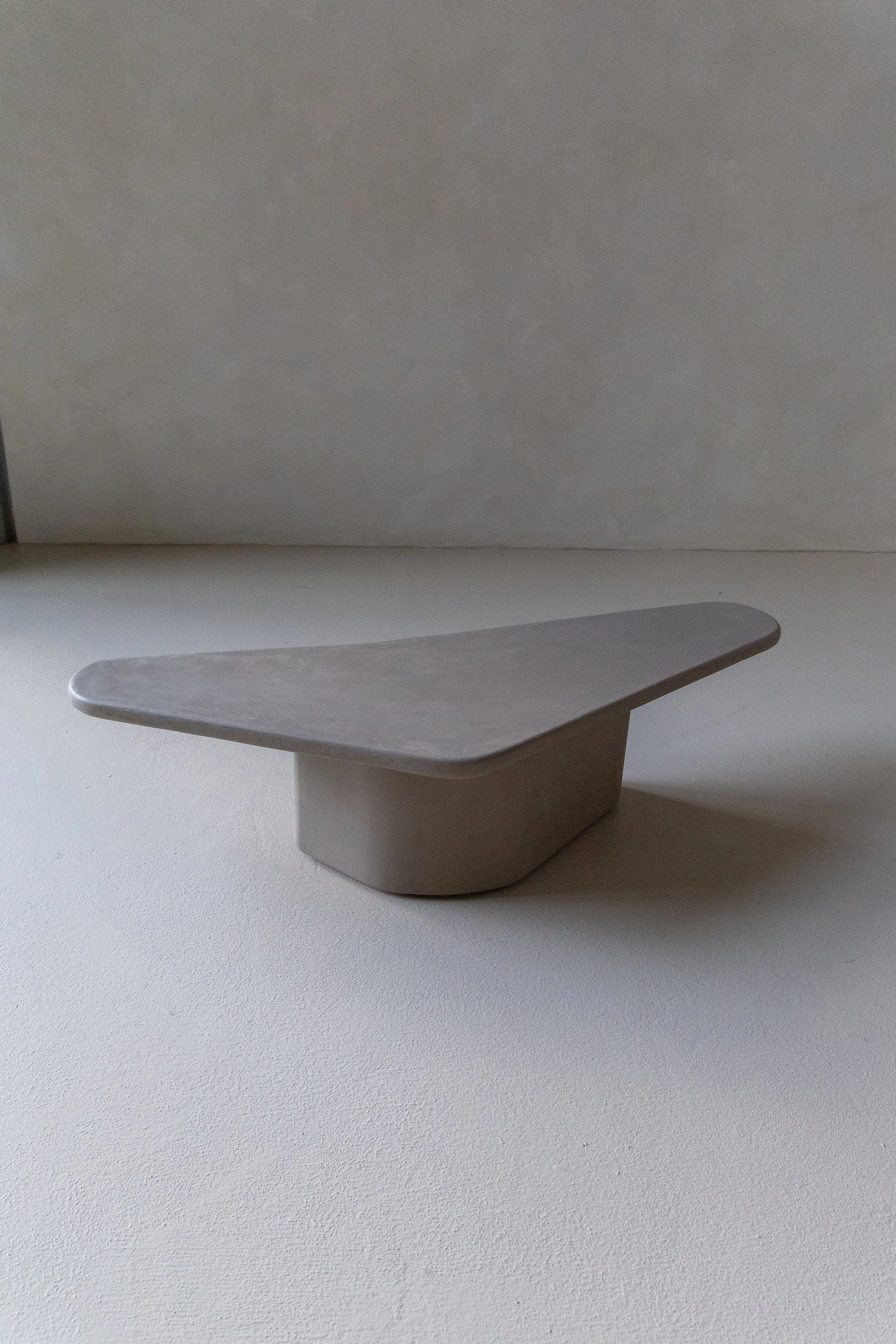 A minimalist, modern, light gray table or pedestal with a geometric shape, placed on a light-colored surface against a plain wall.