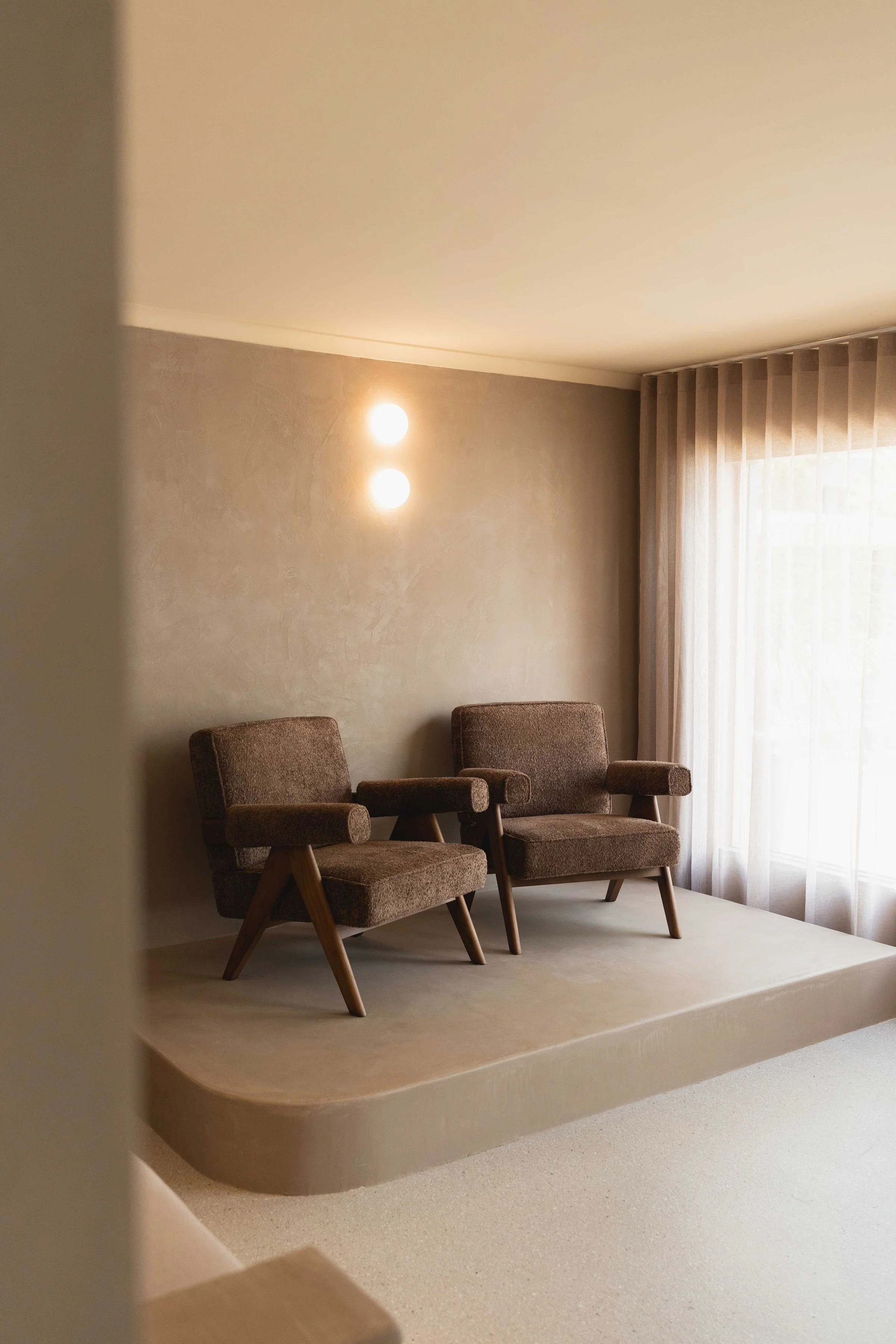 Two brown vintage armchairs on a small stage with beige textured wall and sheer curtains in background.