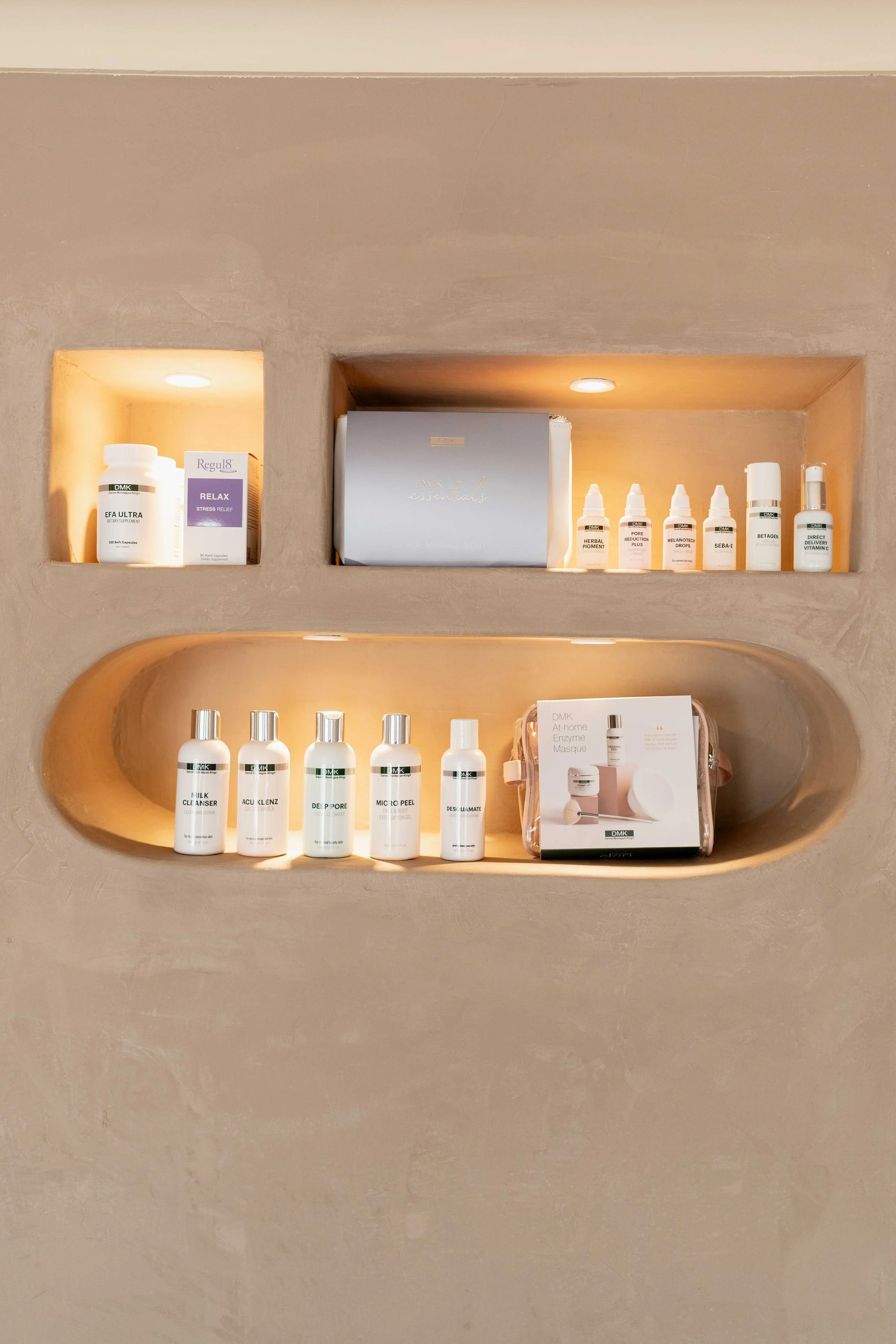 Skincare products arranged on built-in wall shelves, including bottles, boxes, and small containers with a neutral beige background and warm lighting.