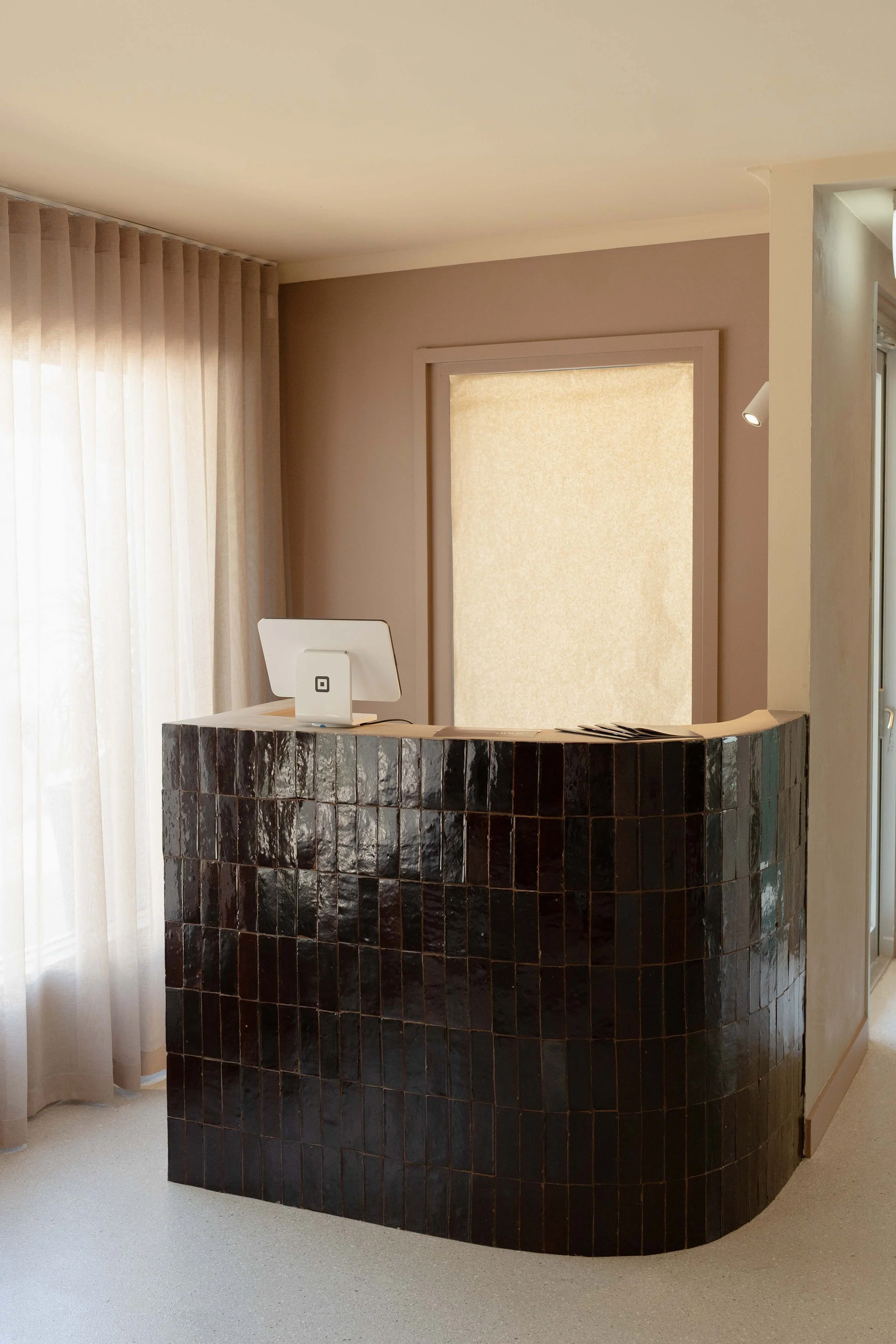 Reception desk with black tiled curved front, a white square-shaped device on top, and a beige curtain and window behind.