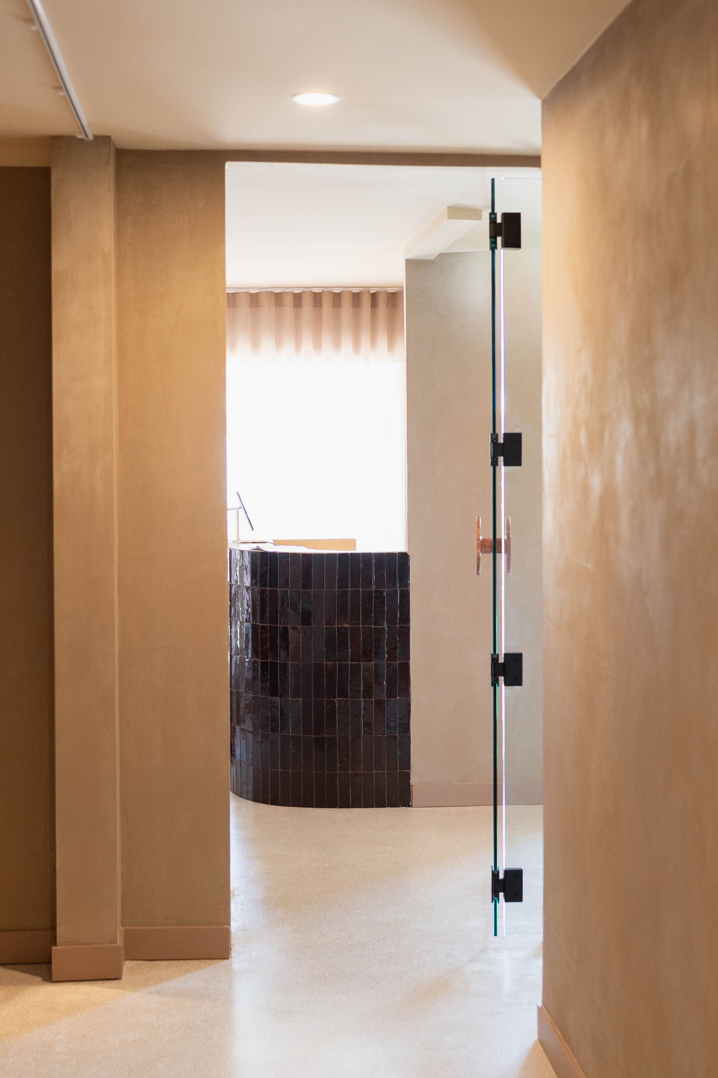 A view through a doorway into a room with a reception desk, illuminated by natural light, with beige walls and a curtain in the background.