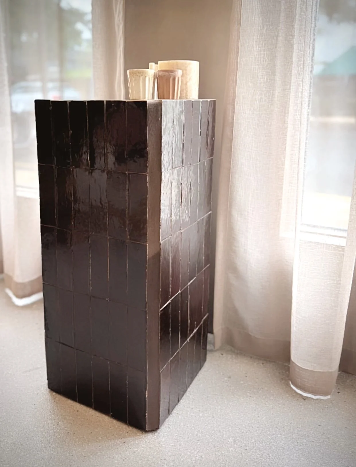 A dark brown tiled pedestal with three candles on top, standing in front of a window with light-colored curtains.