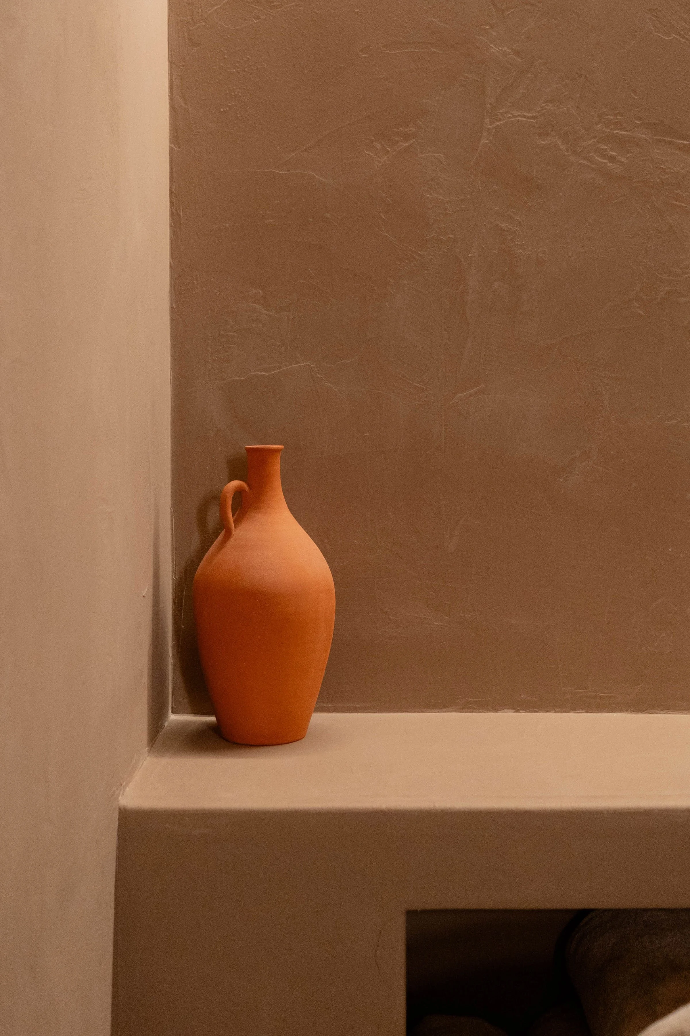 A single small terracotta vase with a handle, placed on a beige ledge against a textured brown wall.
