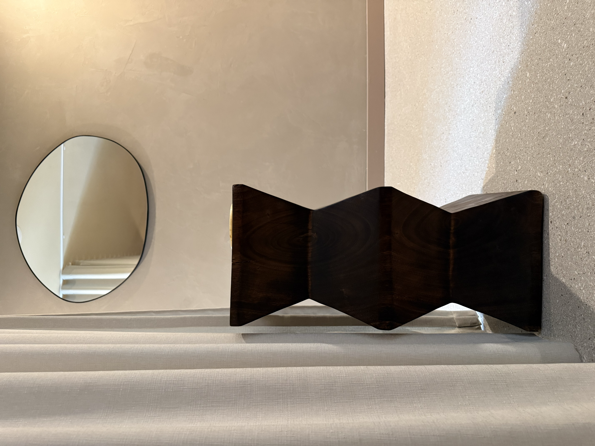 A sculptural wooden stool with an angular, geometric design placed on a carpeted floor, with a wall-mounted circular mirror reflecting part of a room.