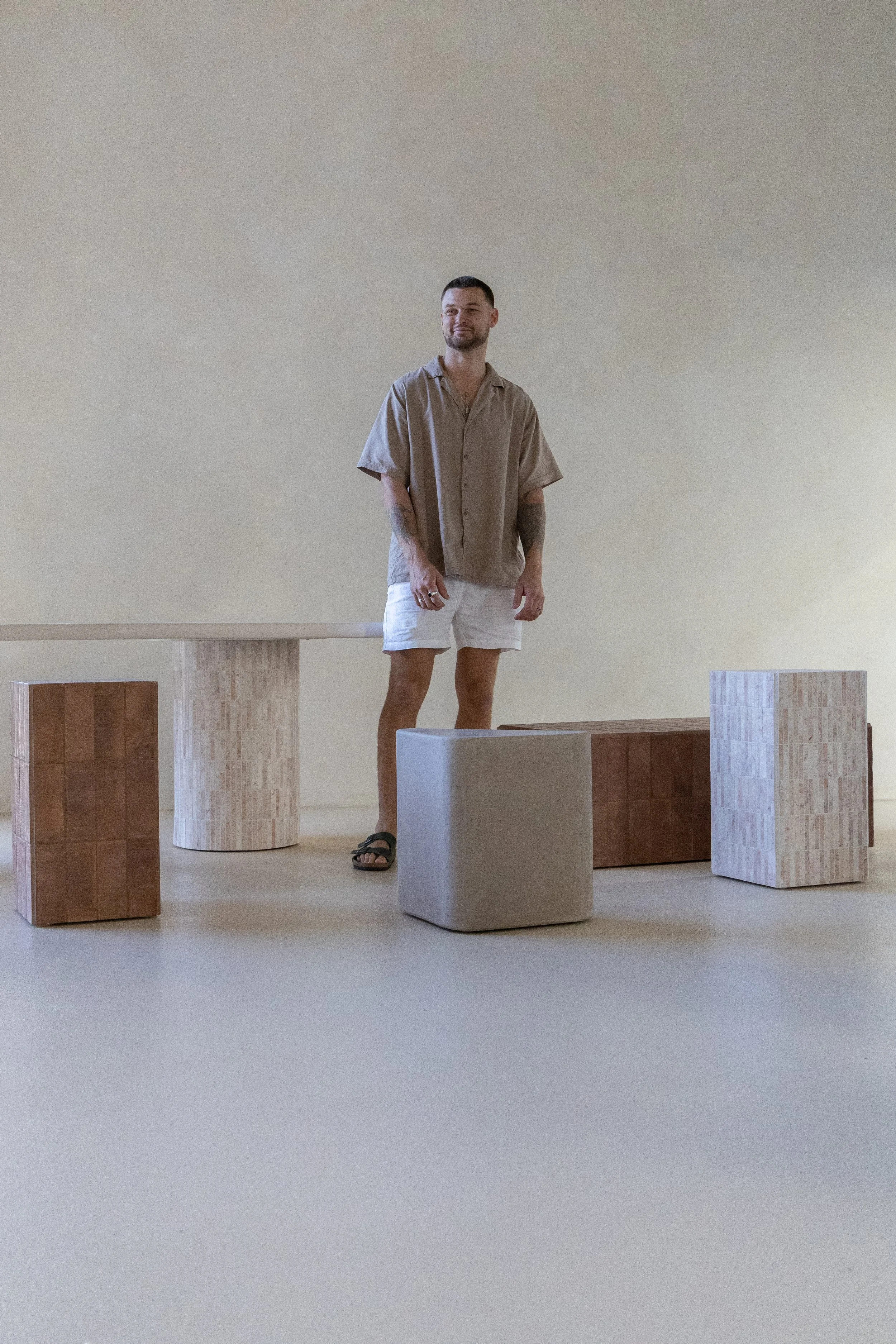 A man standing in a minimalistic room with neutral-colored walls, surrounded by various modern, geometric furniture pieces in earthy tones.