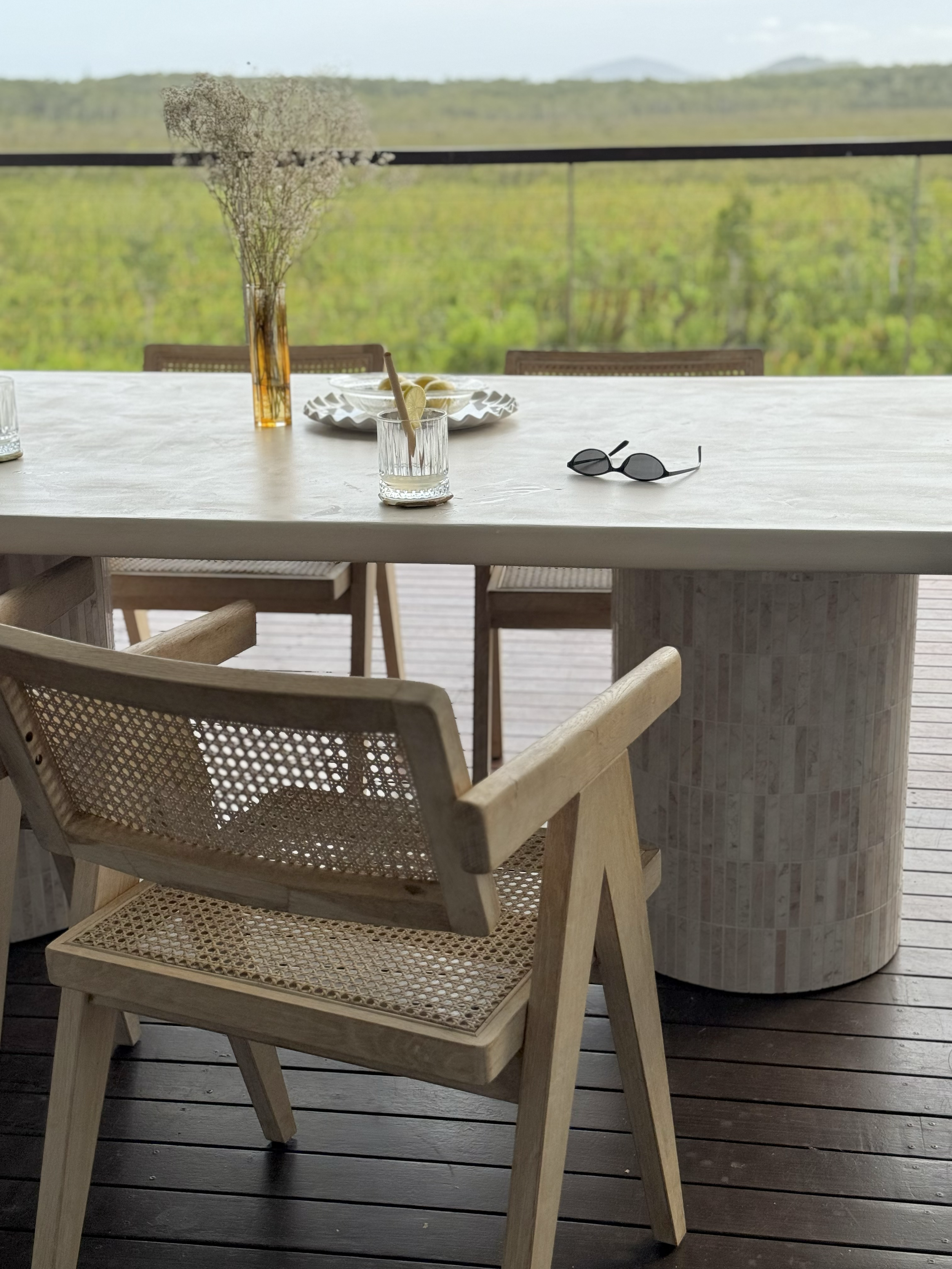 A wooden chair with a woven seat and backrest. In front of a white table with a tall vase of dried flowers, sunglasses, and glasses of lemon water, overlooking a scenic green landscape.