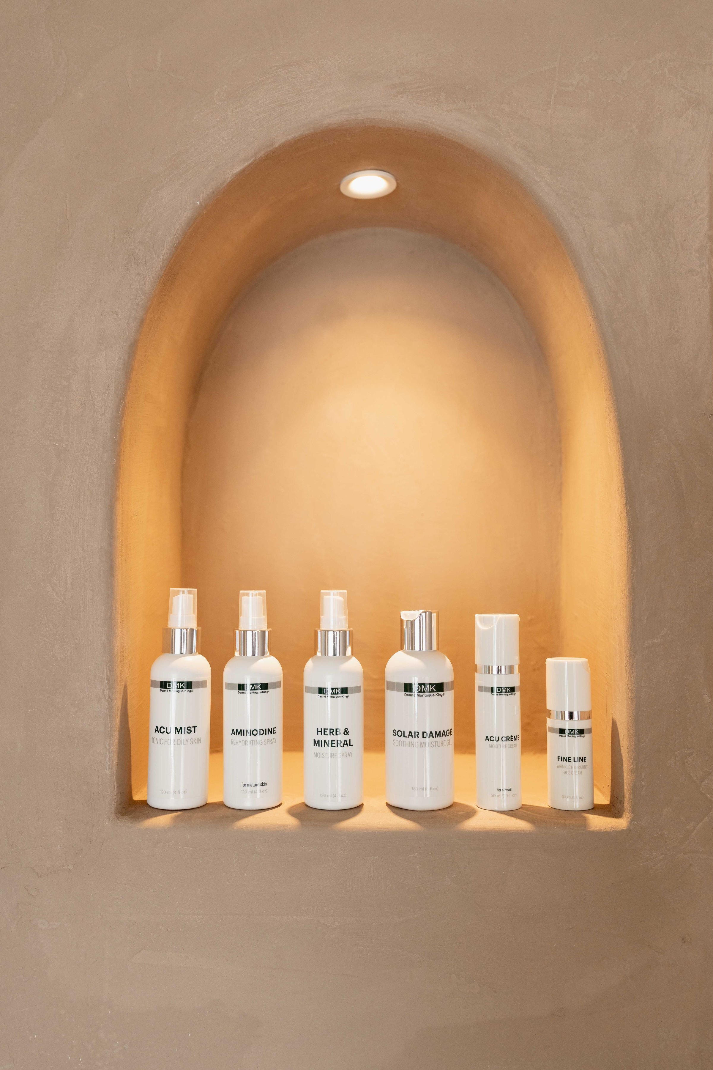 Six white skincare bottles labeled 'Acu Mist,' 'Amino Dine,' 'Here & Mineral,' 'Solar Damage,' 'Acu Creme,' and 'Fine Line,' placed on a built-in shelf within a stucco wall niche illuminated by a small ceiling light.