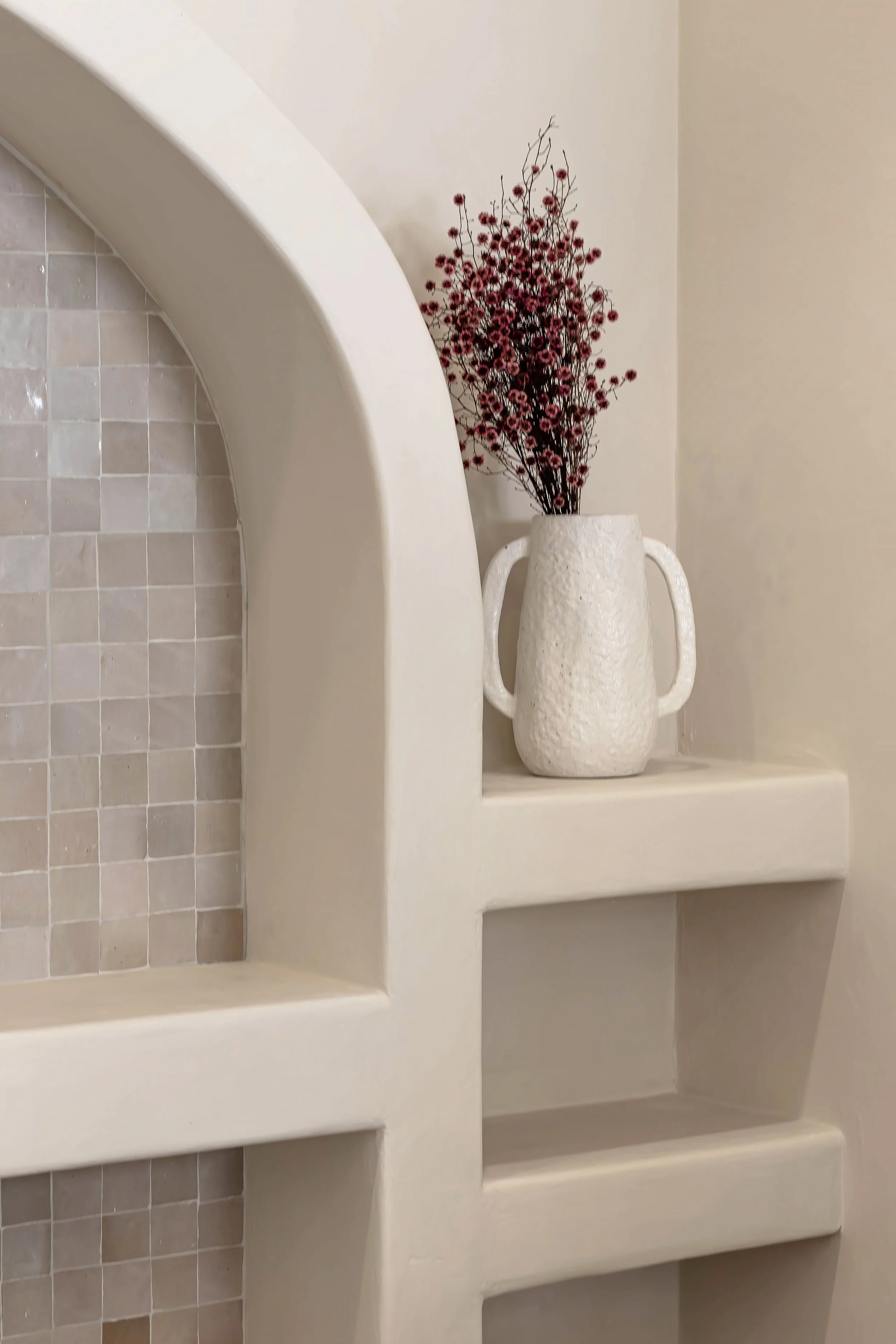 A white textured ceramic vase with two handles contains pink and red dried flowers, placed on a built-in wall shelf with beige walls and tiled section.