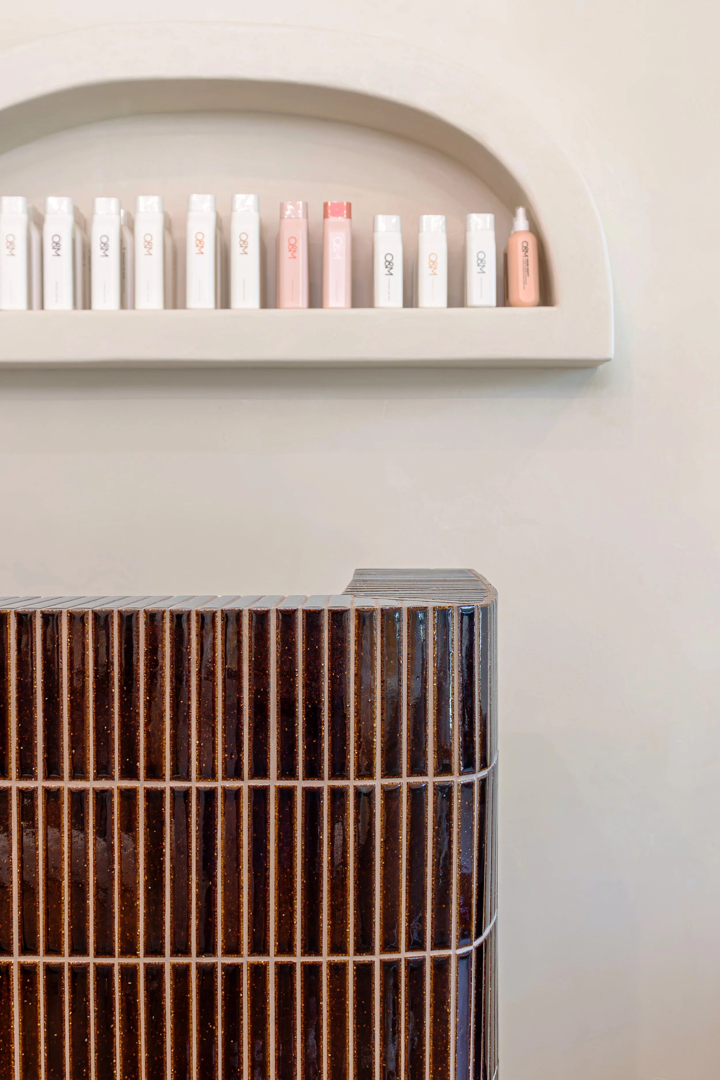 A row of white, pink, and peach cosmetic or skincare bottles on a shelf above a modern, textured, dark brown and black tiled surface.