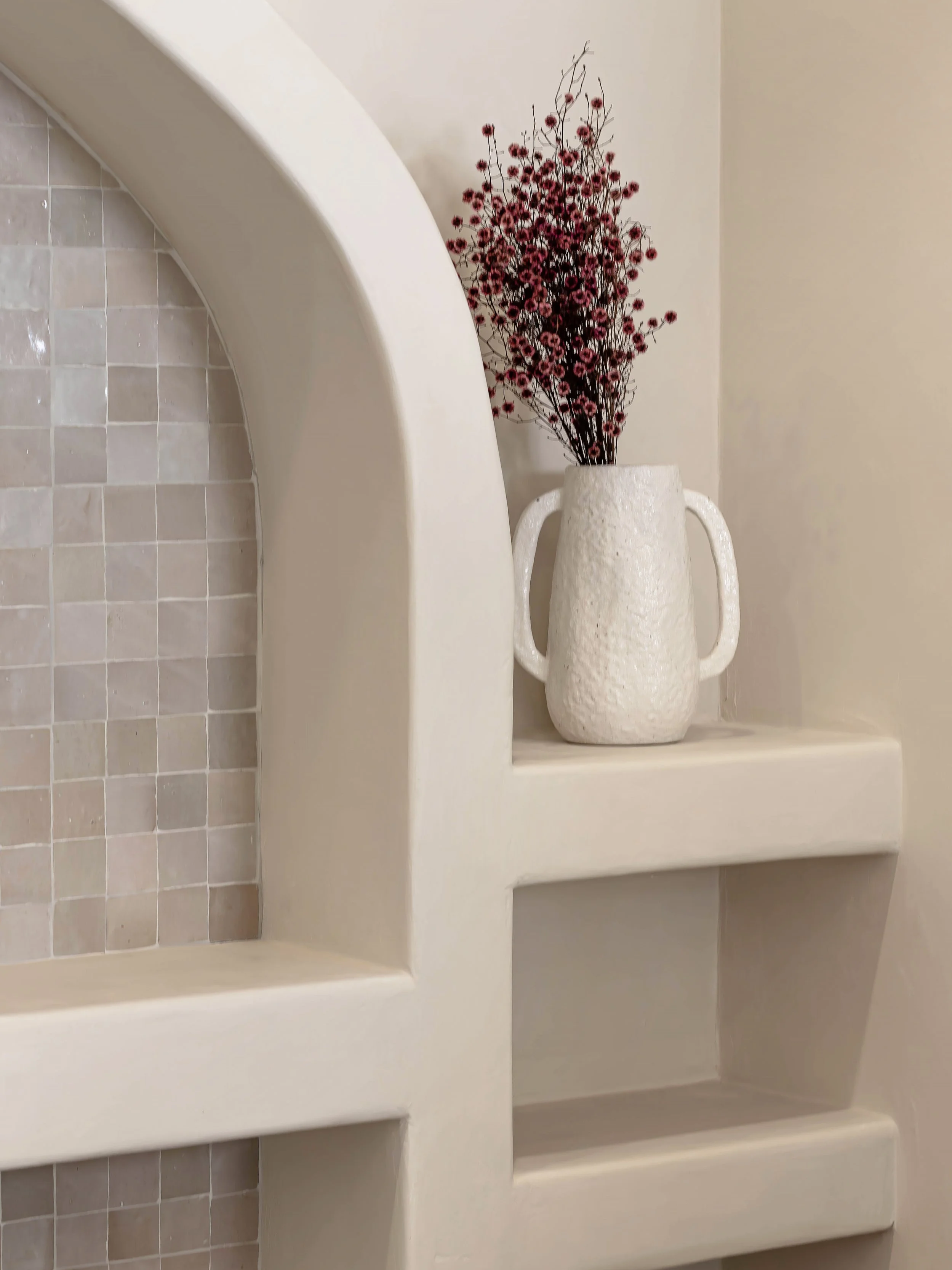 A beige ceramic vase with handles, filled with dark red and pink dried flowers, sits on a ledge in a neutral-colored room with beige walls and a tiled surface.