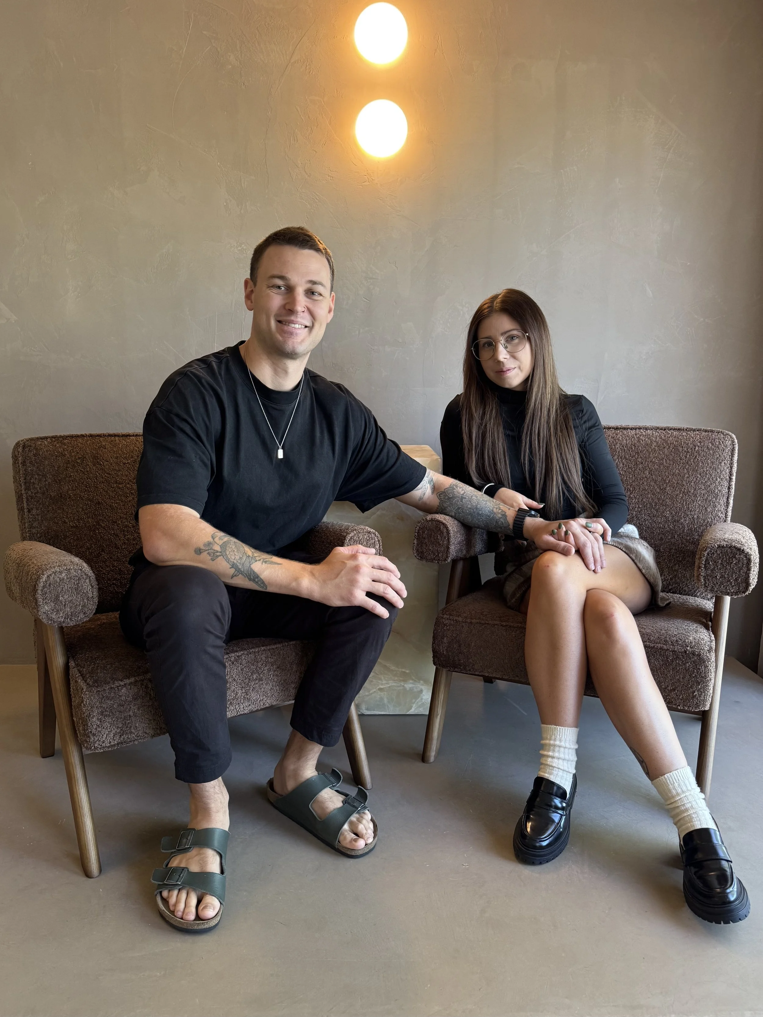 A man and woman sitting on brown armchairs in front of a gray wall with two warm lights overhead, holding hands and smiling.