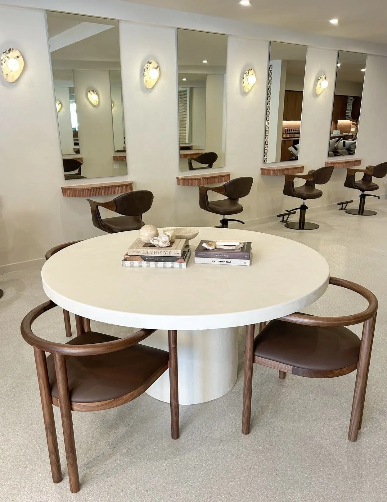 Modern salon or hair studio with a round table and two wooden chairs in the foreground, mirrors and styling chairs along the wall in the background, with soft lighting.