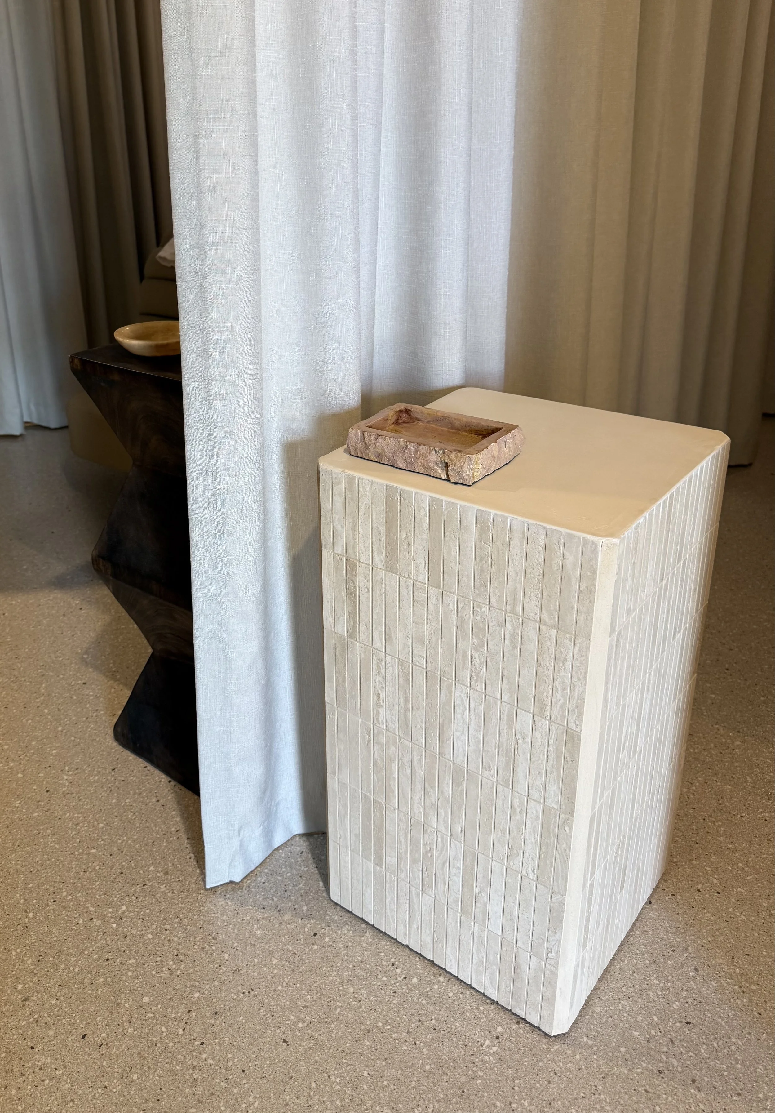 A beige tiled pedestal with a rectangular stone tray on top, positioned next to a light curtain in a room with beige flooring.