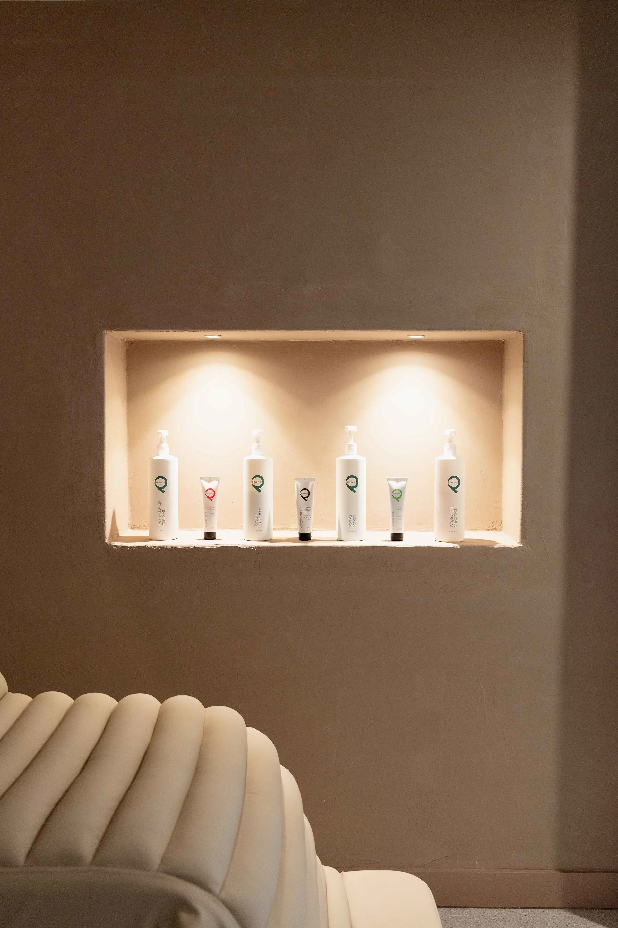 A built-in wall shelf with four illuminated bottles of skincare products, surrounded by white and beige tones, with part of a padded white chair in the foreground.