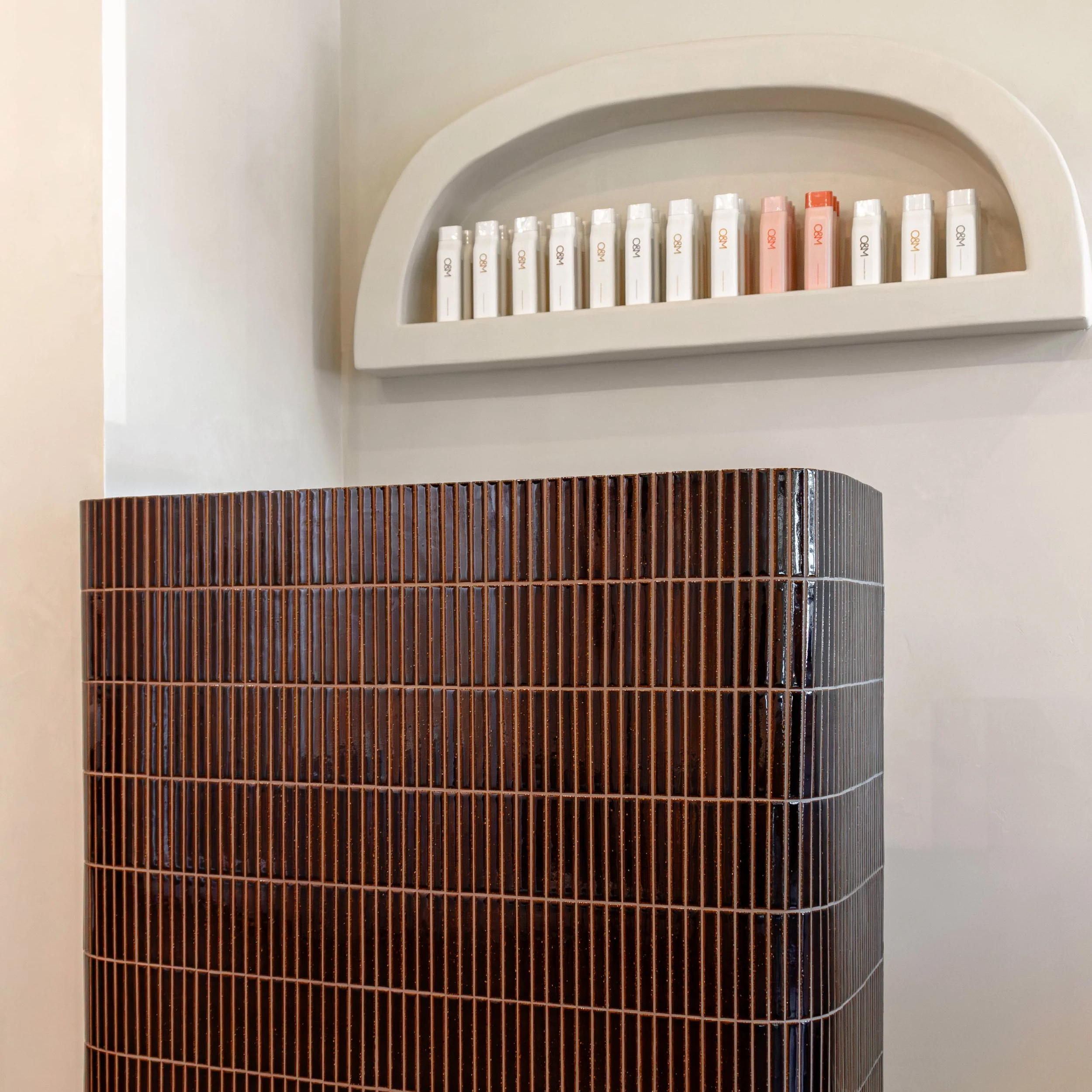 A display of white and pink skincare products on a white, semi-circular shelf, with a brown tiled structure below.