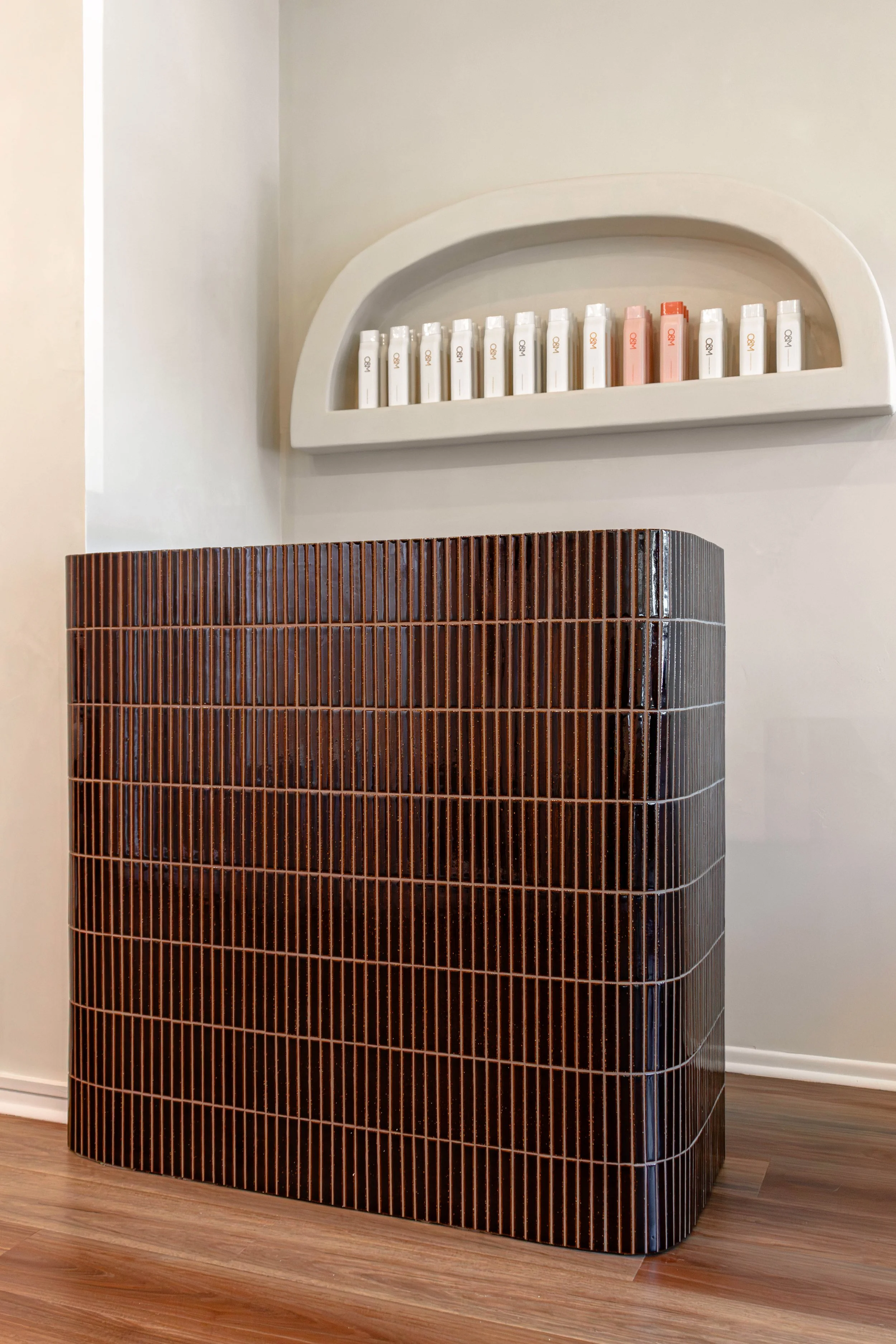 Brown tiled counter with a rounded edge in front of a white wall, a white shelf above with various small bottles and containers, dark hardwood floor.