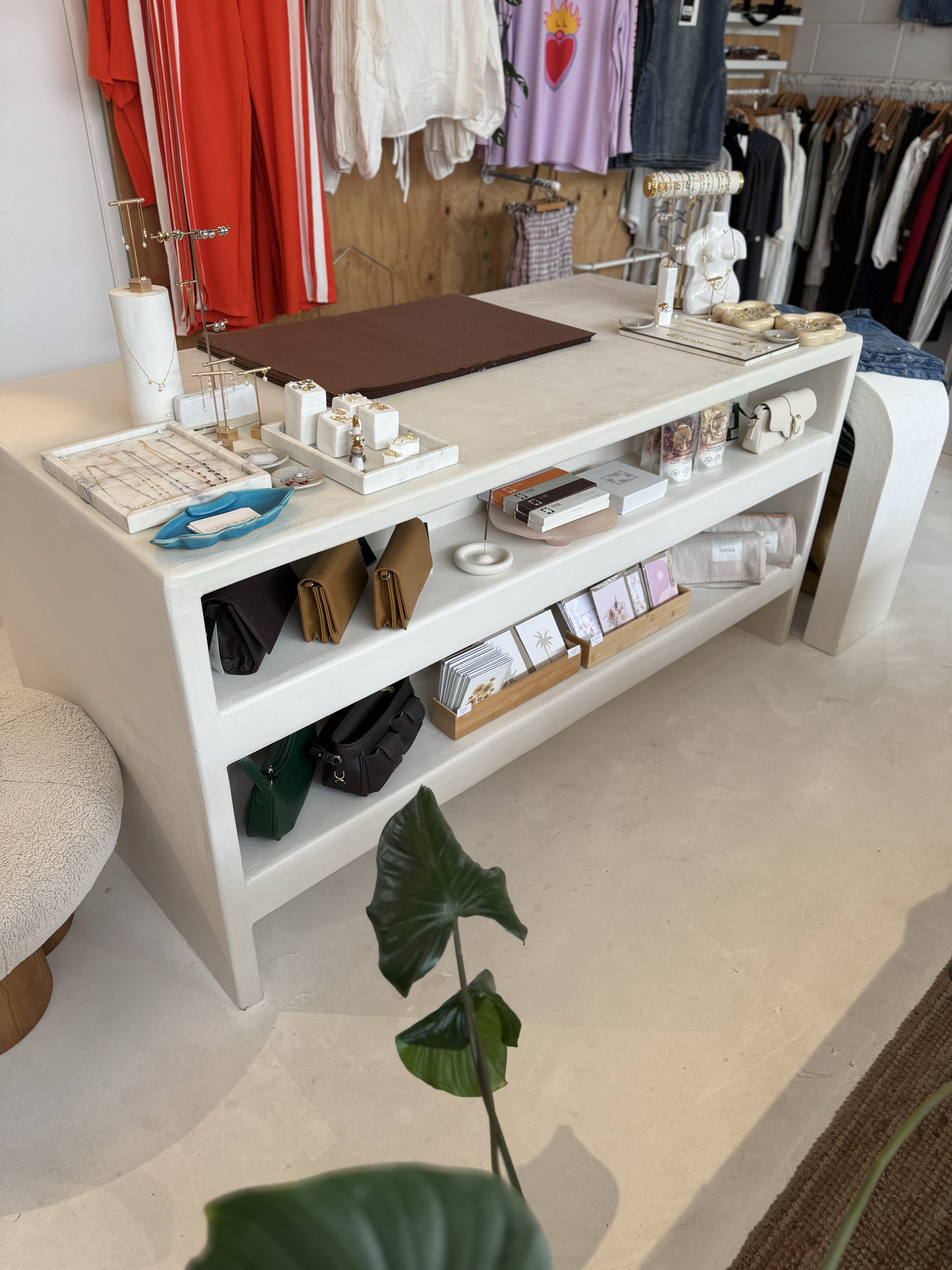 Display table in a boutique with jewelry, wallets, candles, and small accessories, surrounded by clothing racks and a plant in the foreground.