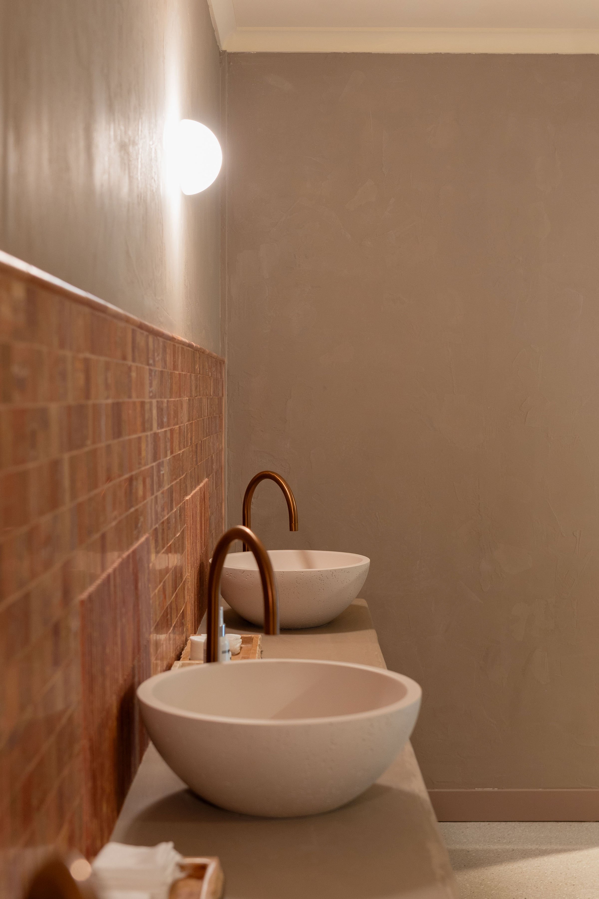 A row of two modern vessel sinks with copper faucets on a beige countertop in a public restroom with a textured wall and a wall-mounted light fixture.