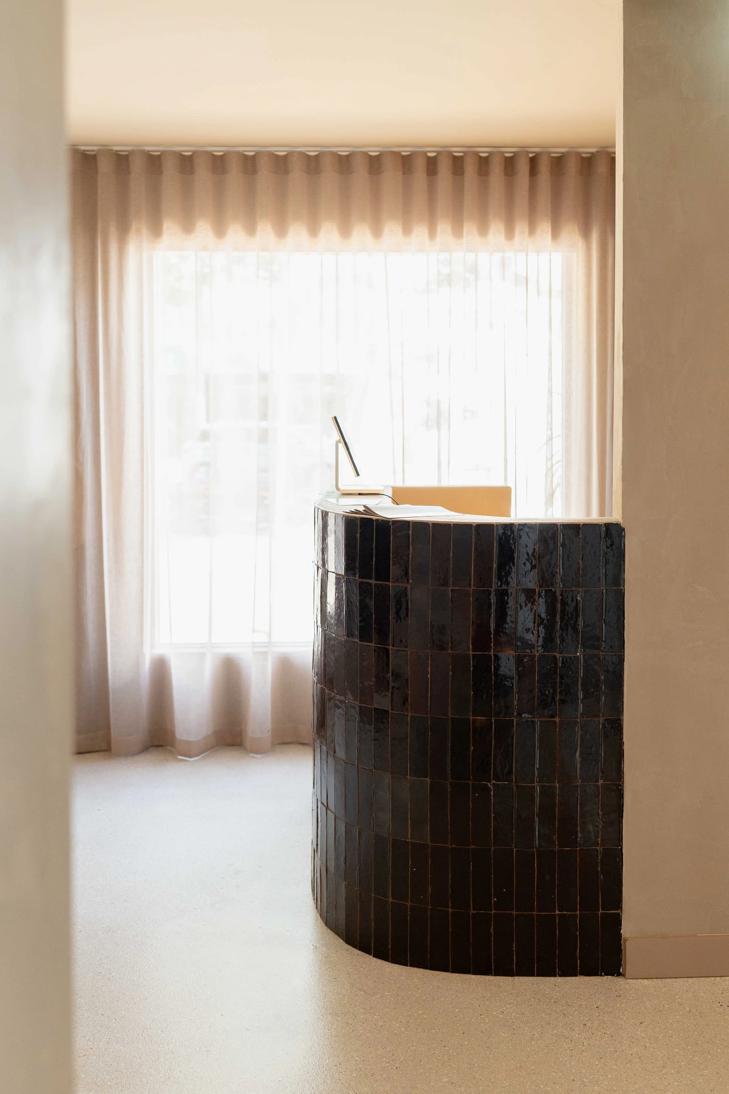 A reception desk with black tiles in front of a large window with sheer curtains, and a computer on the desk.