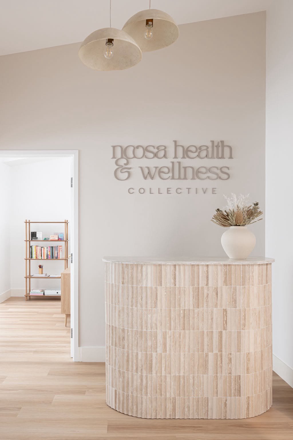 Reception area of Ncosa Health & Wellness Collective with a curved beige tiled counter and a beige vase with dried plants on top. Light wood flooring and minimalist decor.