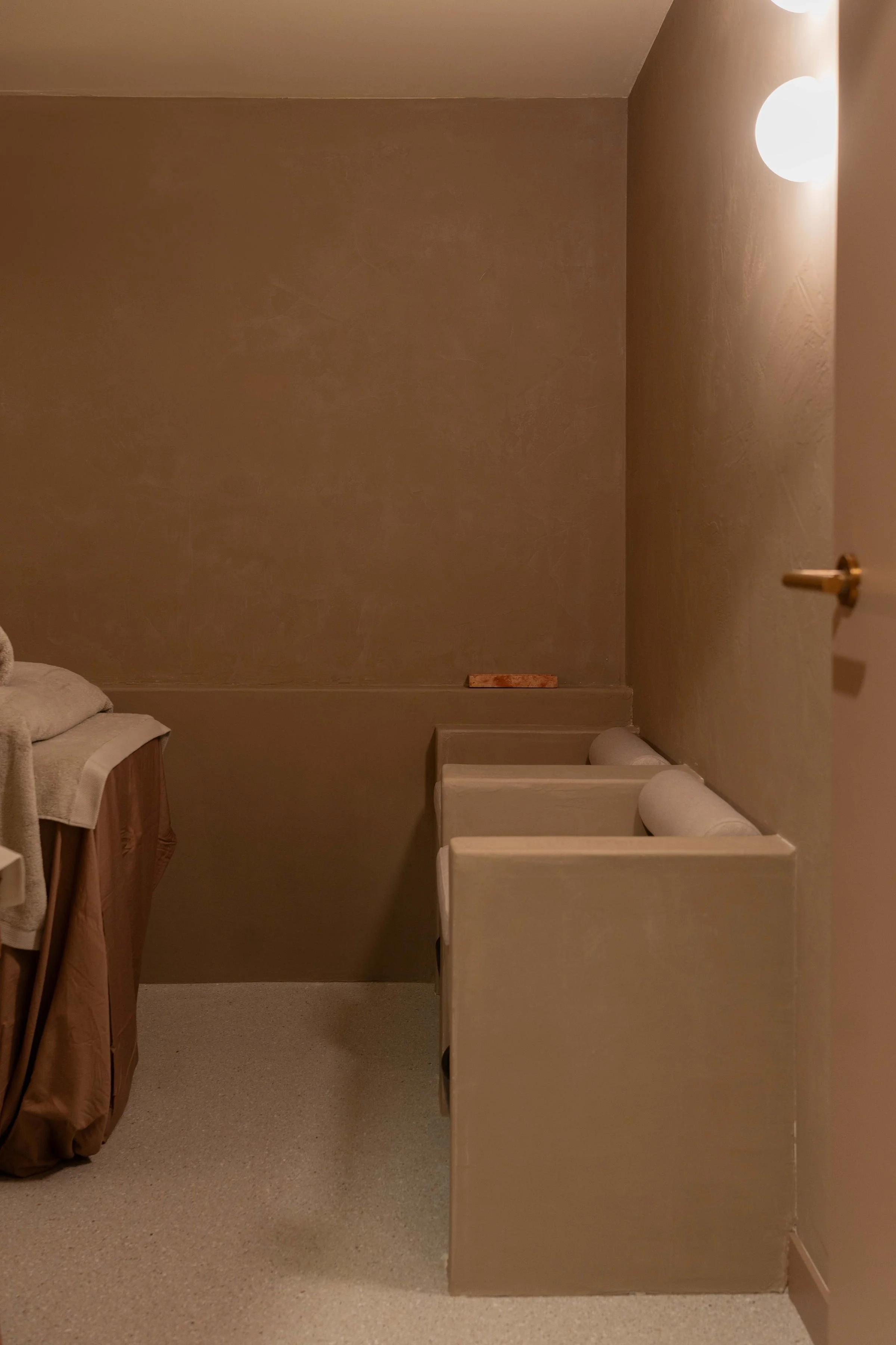 Inside a room with beige walls and carpeted floor, three beige cubicles or machines with cylindrical rollers on top are aligned against the right wall, and a bed with rolled towels is partially visible to the left.