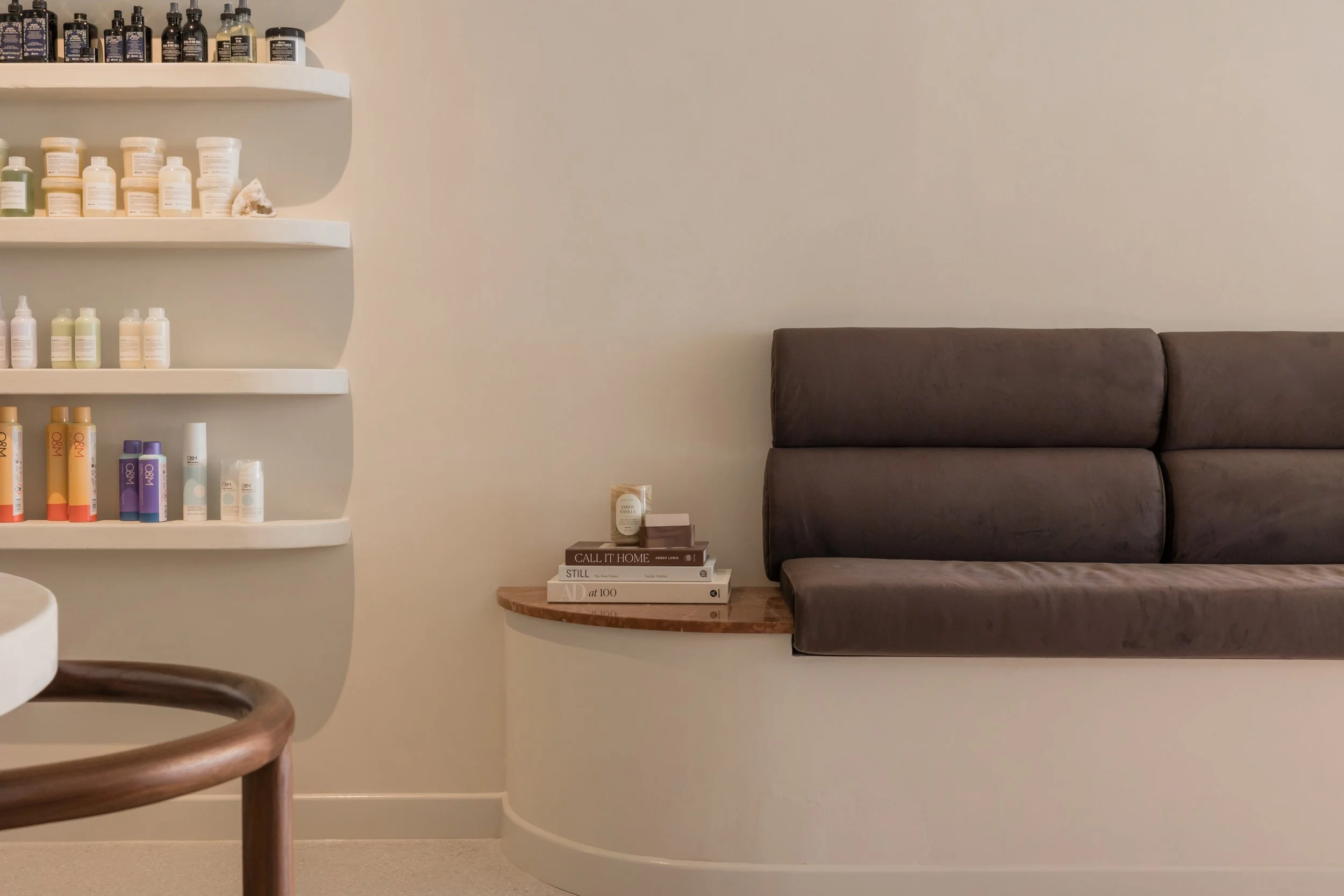 Wall-mounted shelves with bottles and containers, a small stack of books on a wood ledge, and a padded dark gray bench in a room with a neutral background.
