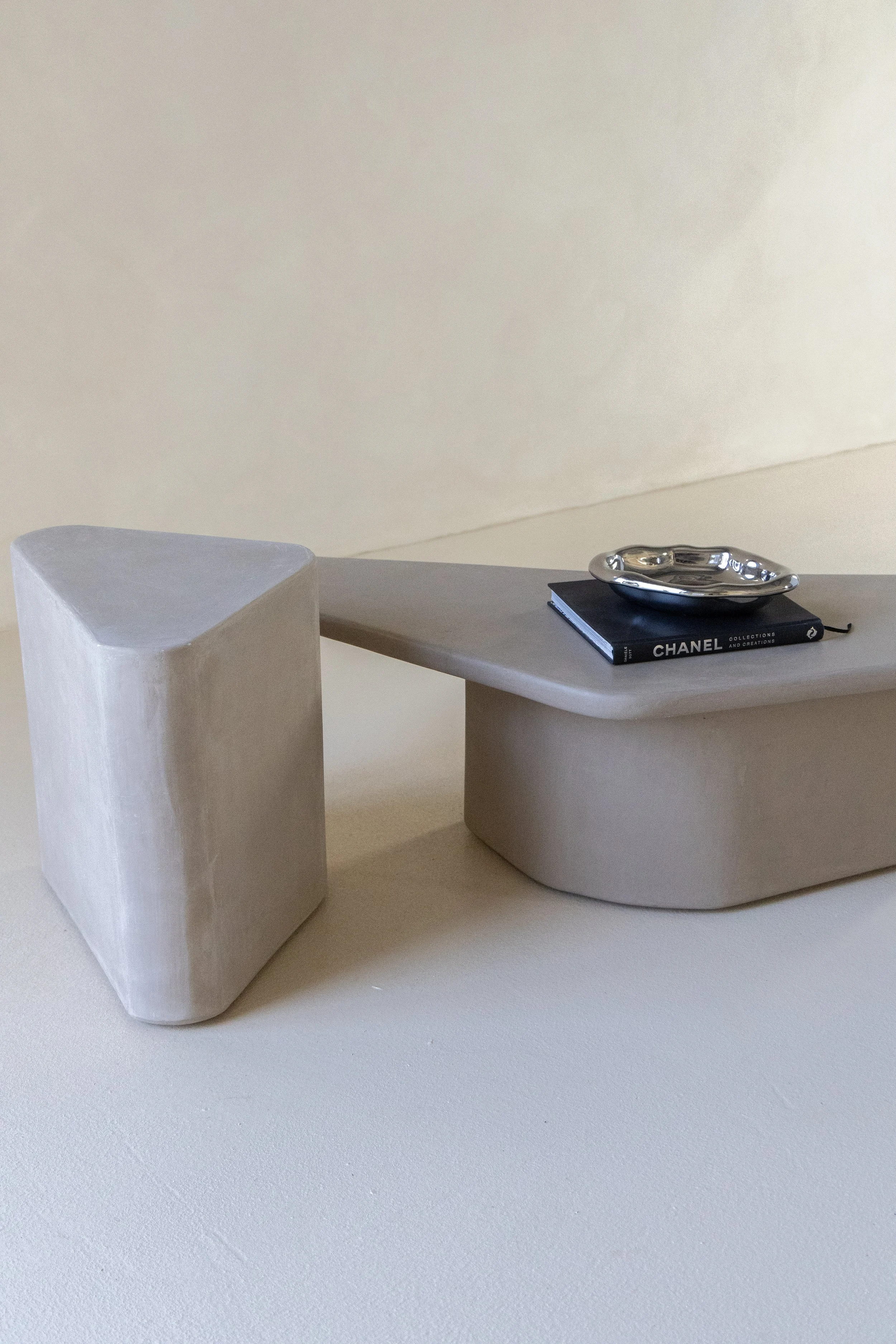 Minimalist decor with a geometric white sculpture, a gray tray, a black Chanel notebook, and a silver dish on top of the notebook
