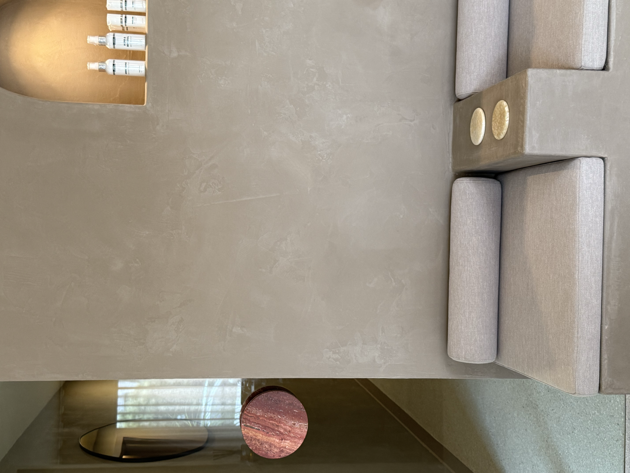 A beige sectional sofa with rectangular and cylindrical cushions, placed against a beige wall with a textured finish. Two small round books or decorative plates in gold are on the armrest. In the background, frosted curtains and a large mirror are vi