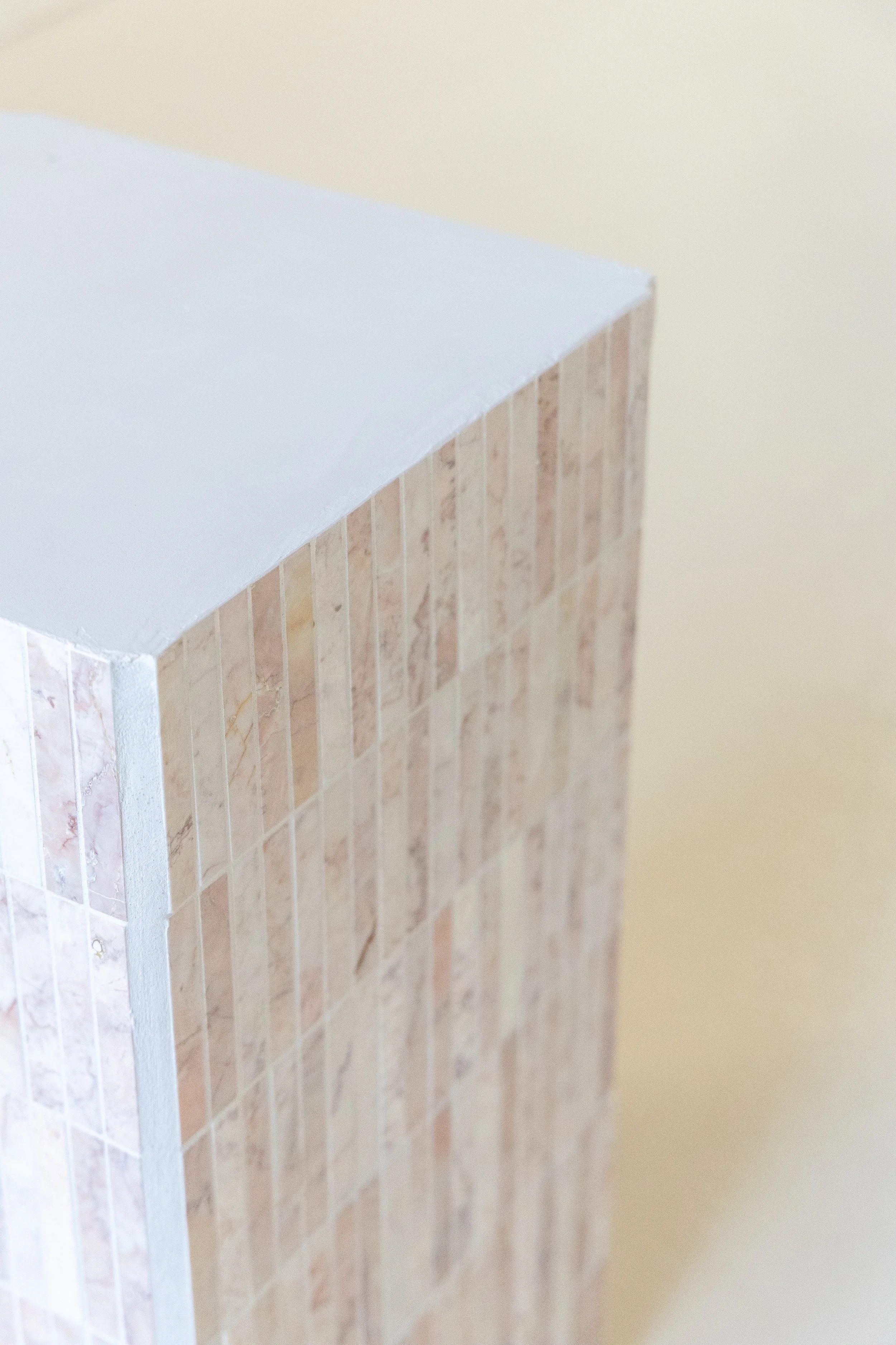 Close-up of a corner of a piece of furniture, showing a white top surface and a side covered with small beige tiles, against a cream-colored wall background.