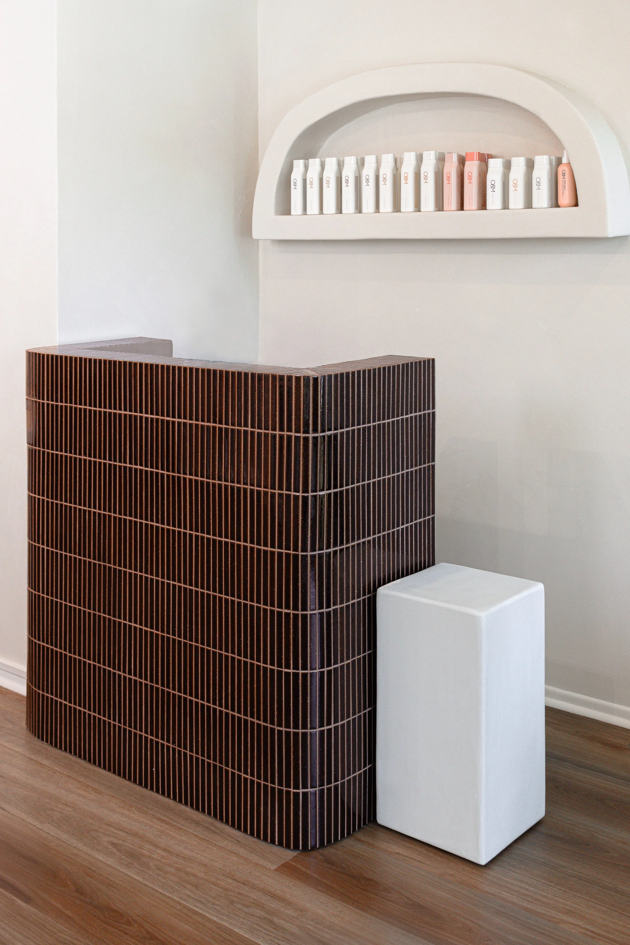 A modern interior with a brown tiled counter, a white cube-shaped stool, and a white wall-mounted shelf with bottles.