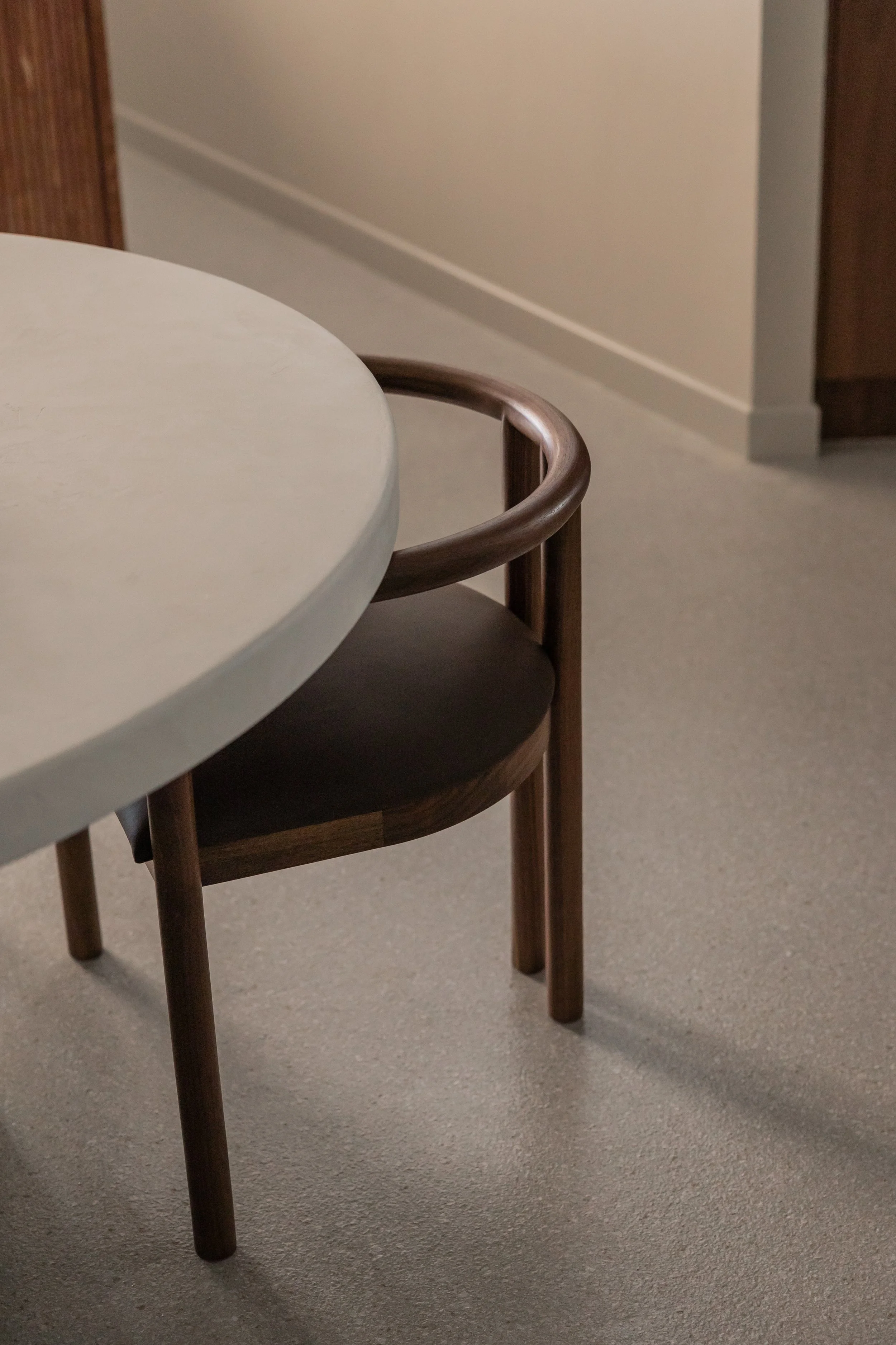 A partial view of a round, light-colored table with a dark wooden chair next to it, situated on a beige carpeted floor in a room with plain walls.