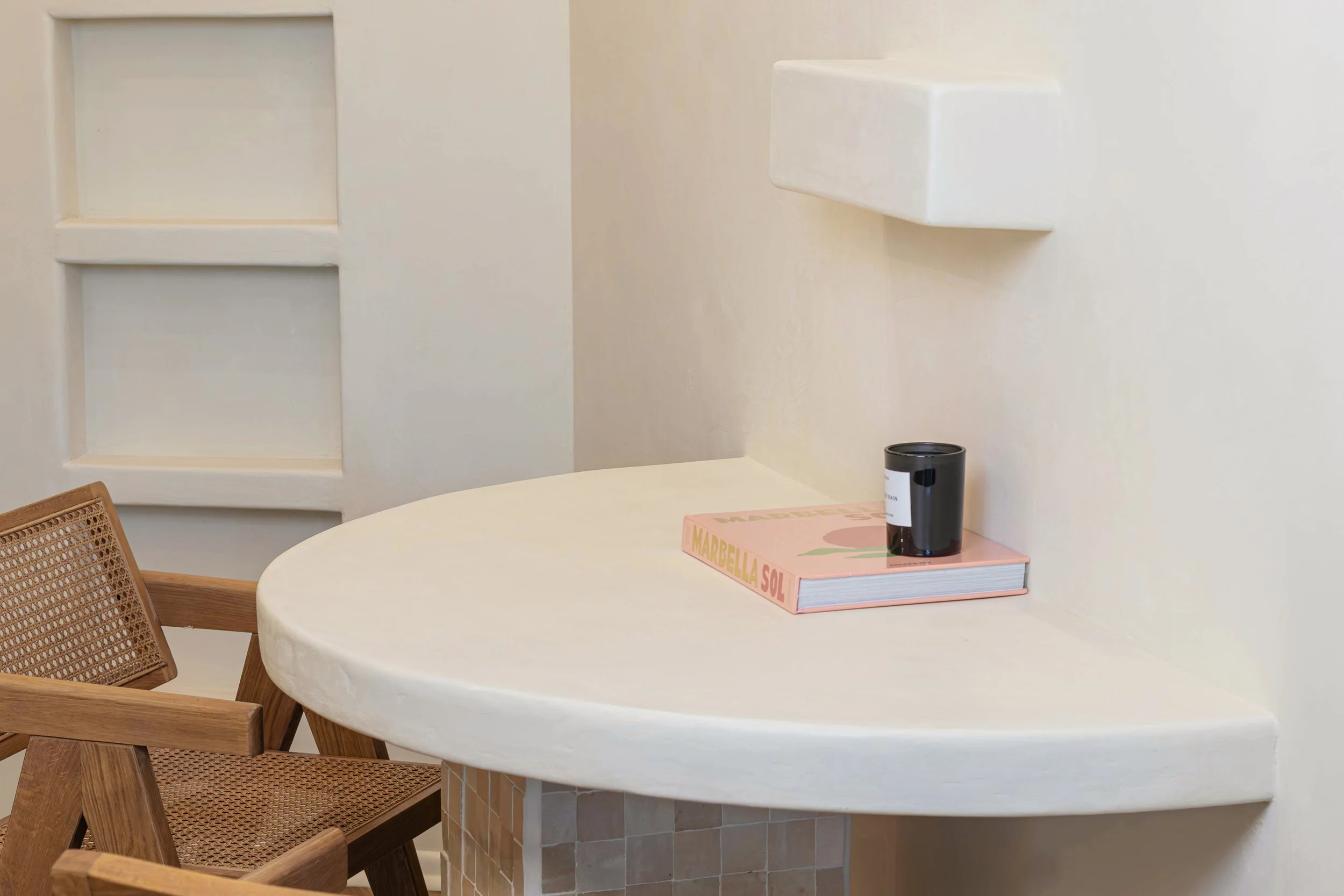 A white, rounded table with a pink book titled 'Marbella Sol' and a black cup on top. There is a wooden chair with a woven seat next to the table, set against a light-colored wall with built-in shelves.